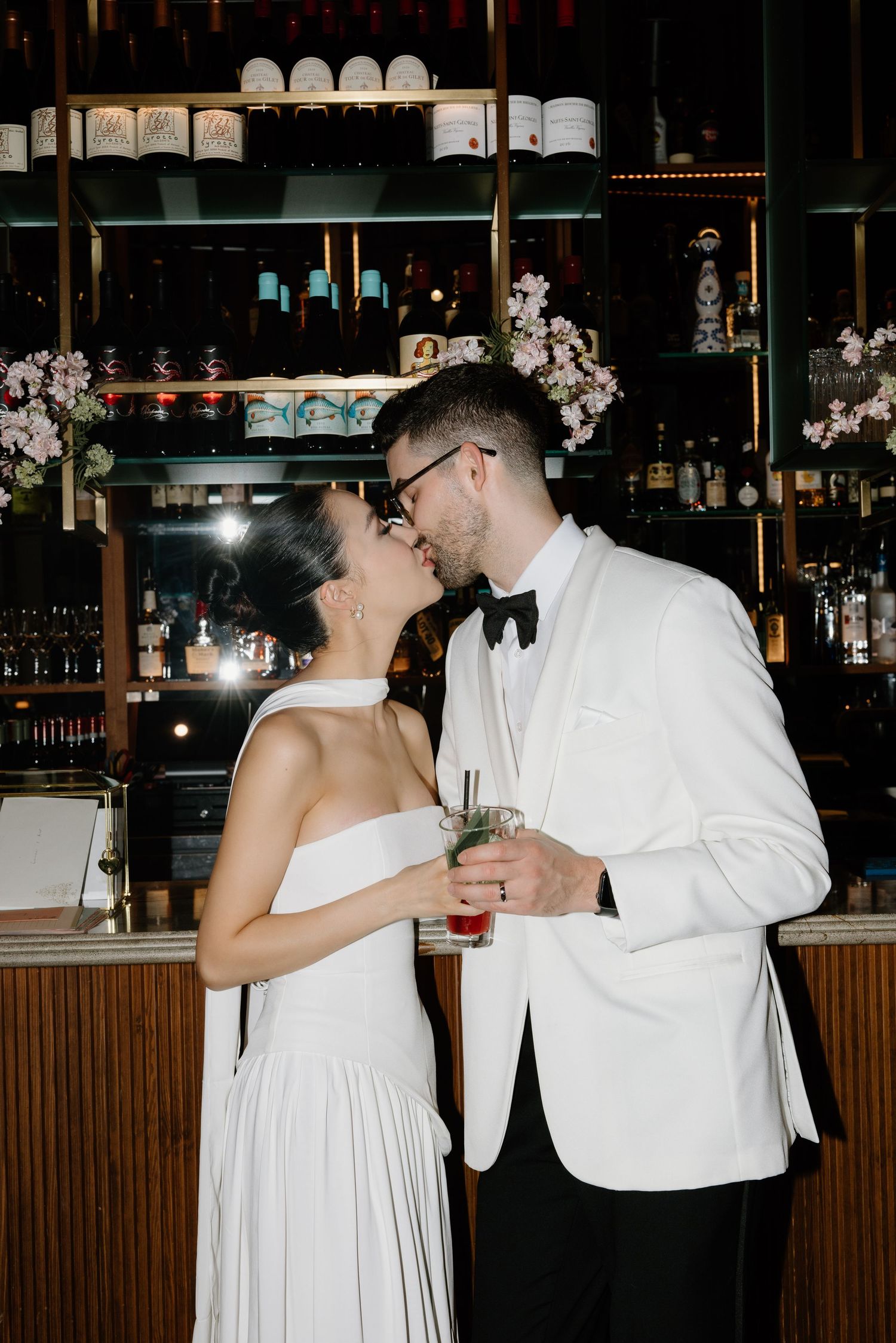 Well-dressed couple in white evening wear embrace while enjoying drinks at a stylish bar with floral decor.