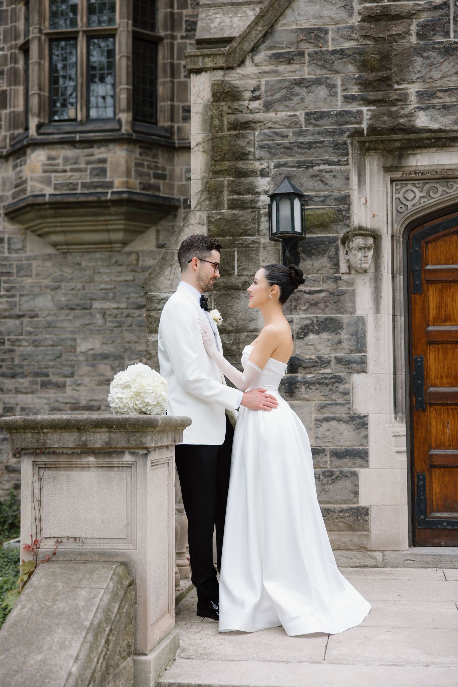 A couple in elegant white wedding attire embrace on stone steps of a historic building with gothic architecture.