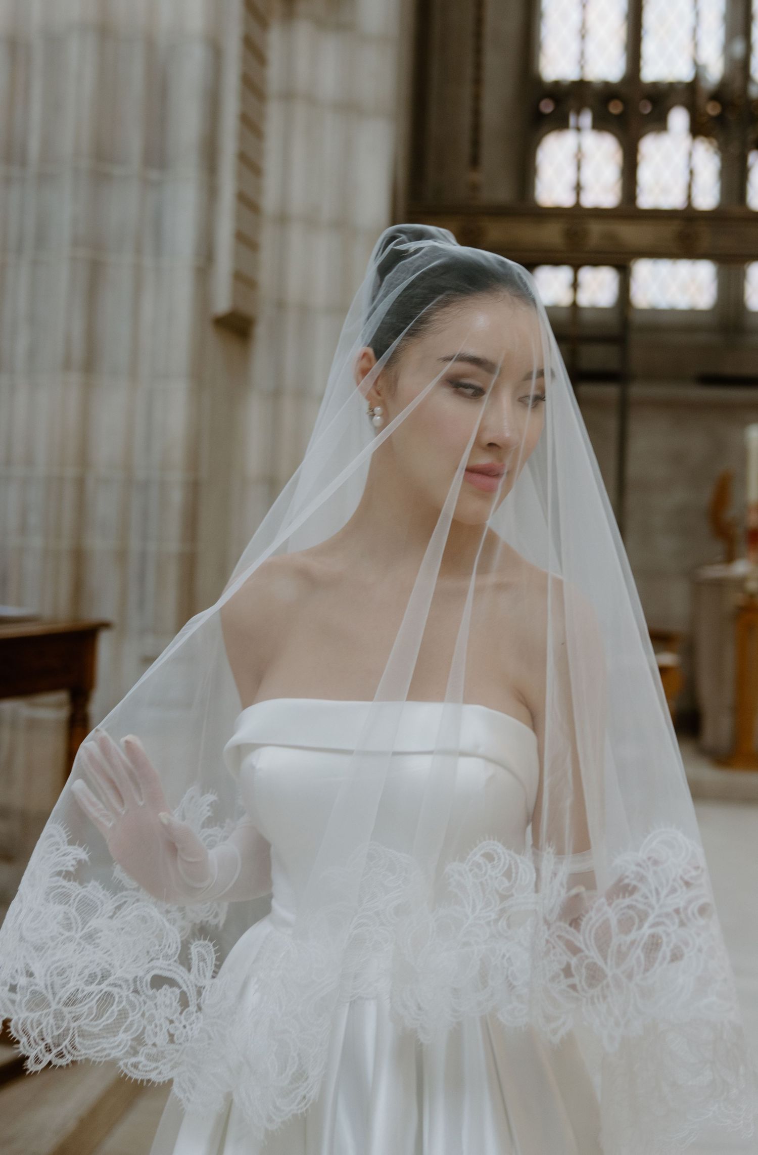 A bride in an elegant white strapless wedding gown with a delicate lace-trimmed veil stands in a grand cathedral setting.