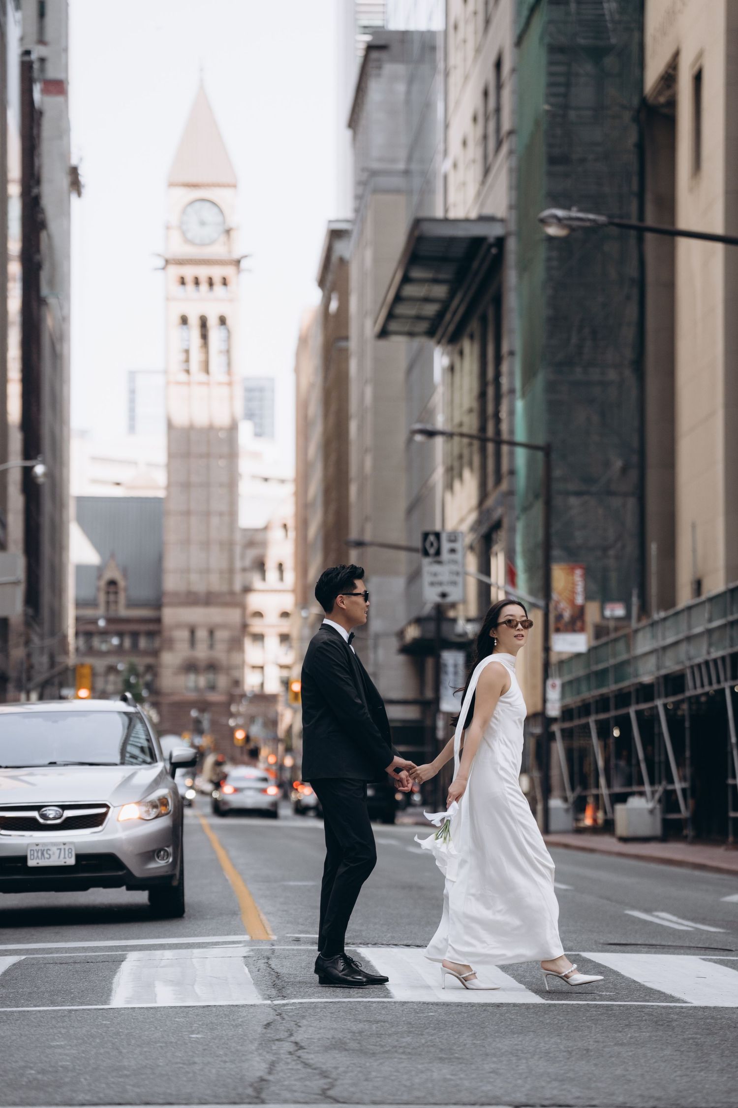 Newlyweds holding hands while crossing city street with historic clock tower and urban architecture in background.