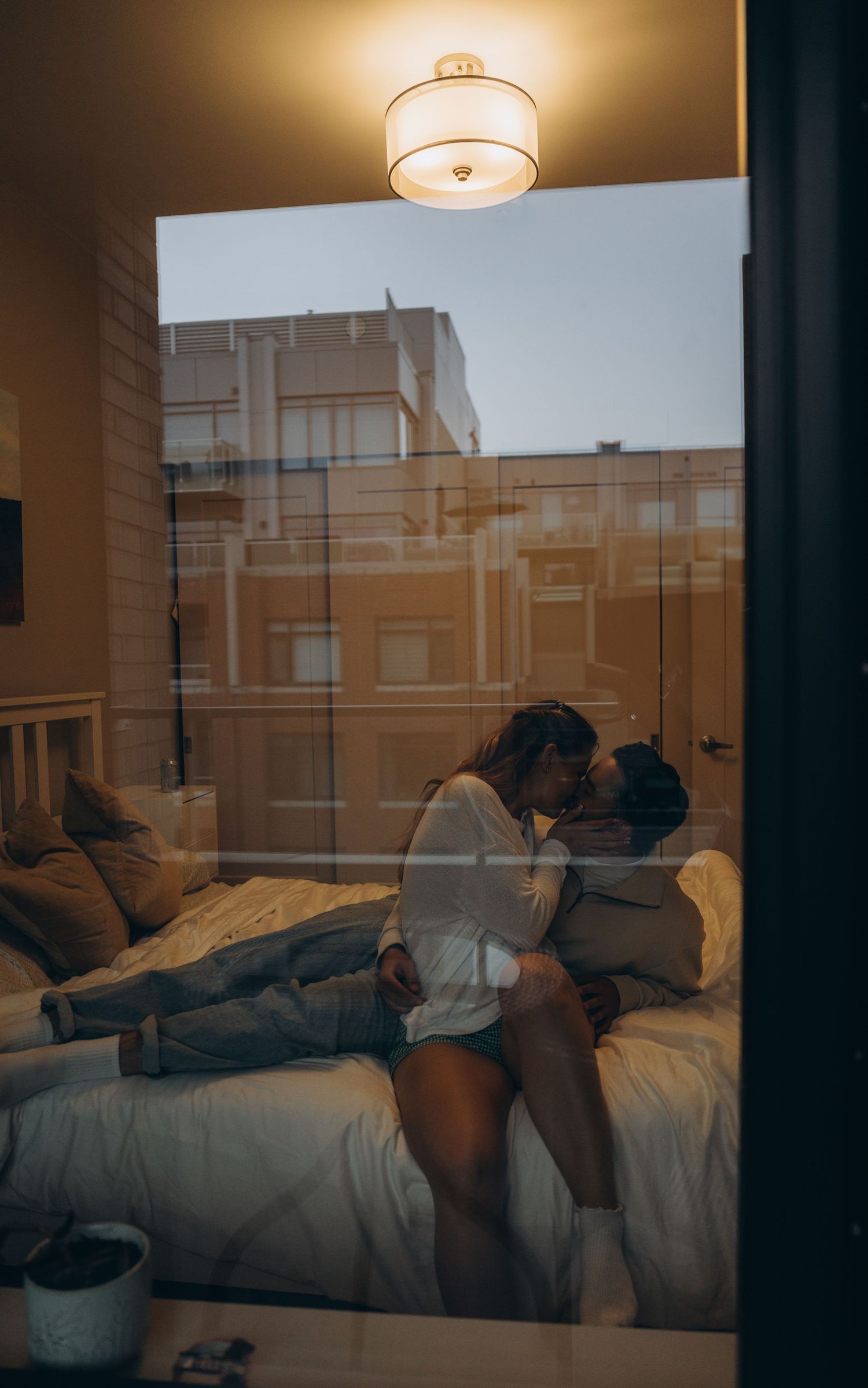 Couple relaxing comfortably together on a bed in a warmly lit bedroom with windows reflecting sunset light.