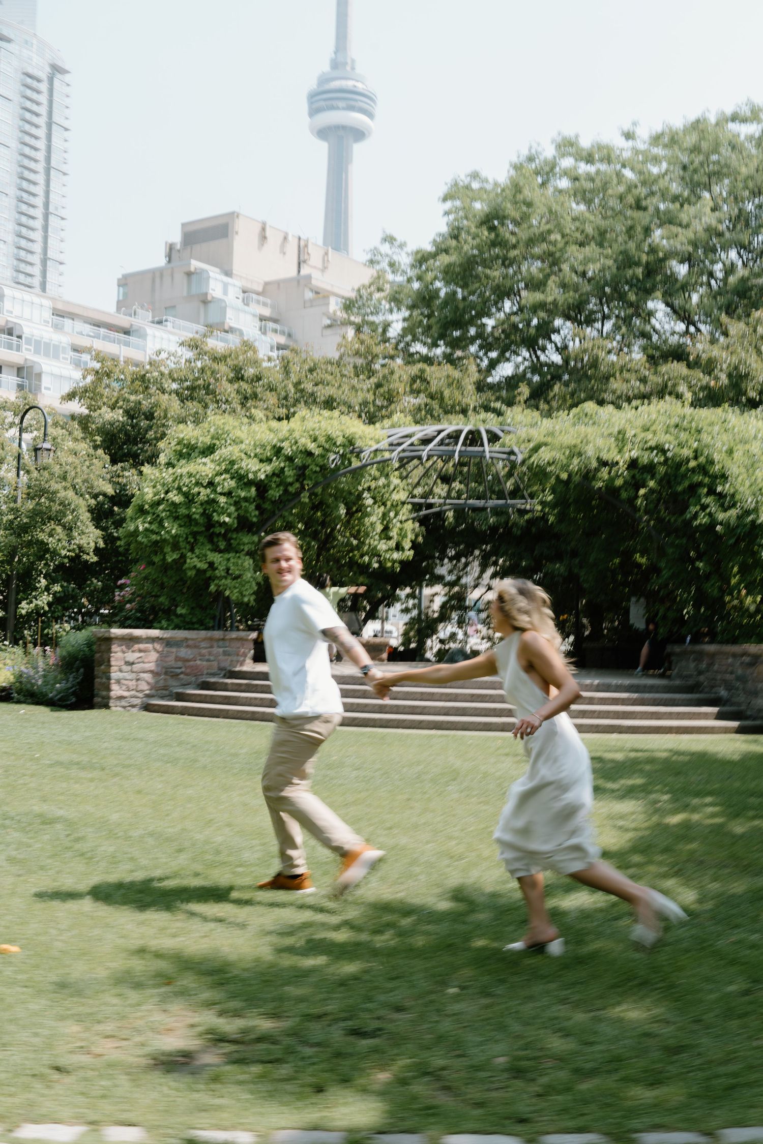 Two people run playfully on a green lawn with the iconic CN Tower visible in the Toronto skyline background.