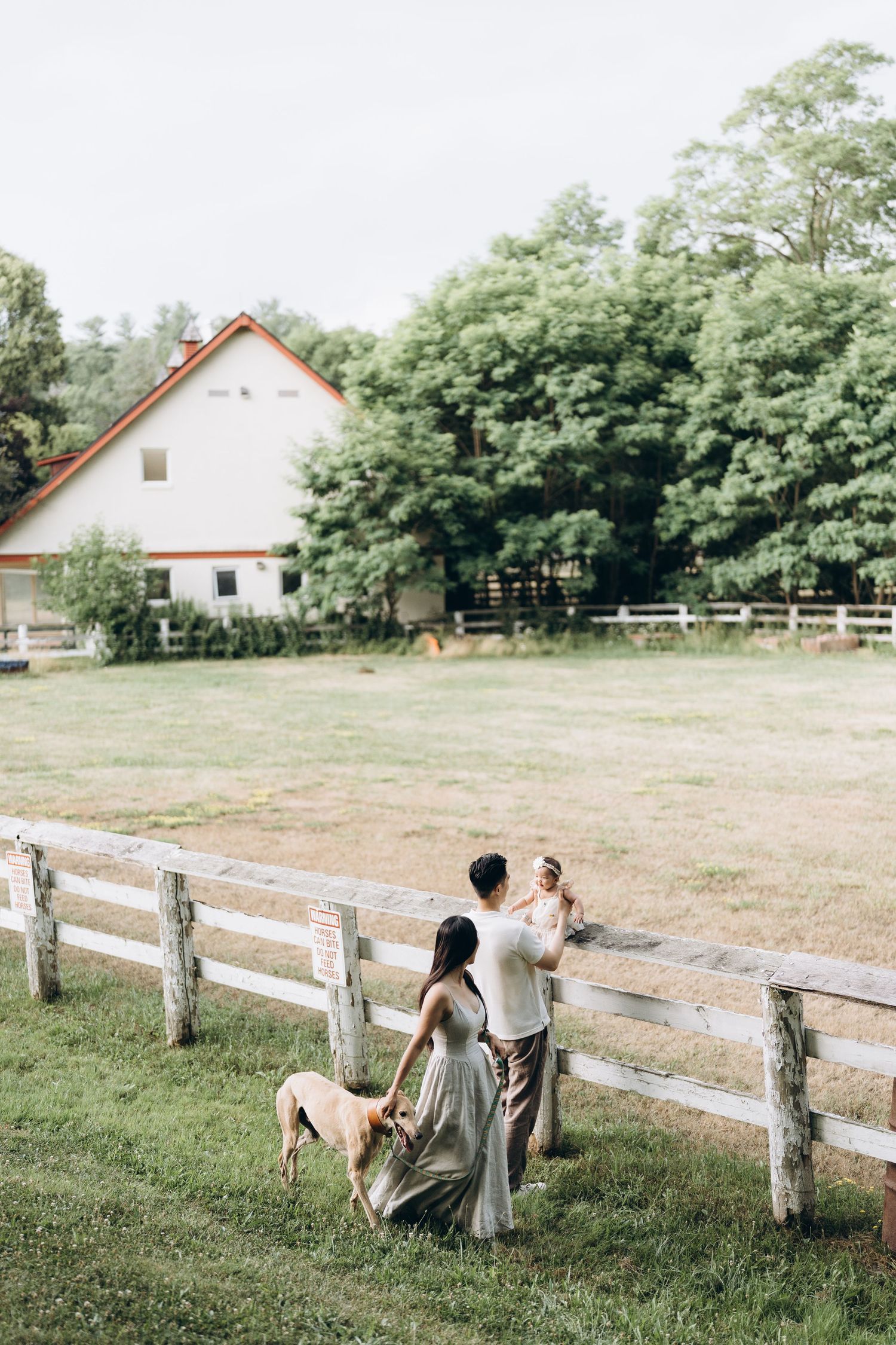 Couple and dog standing by white wooden fence at rustic farm with barn and trees in background.