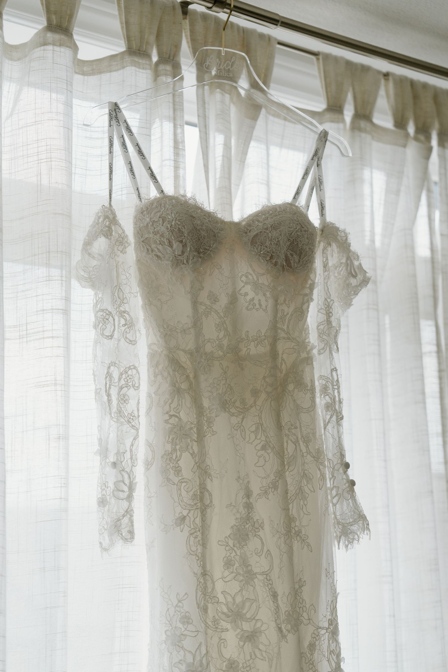 Several vintage white lace wedding dresses hanging on display against a light-filled window with sheer curtains.