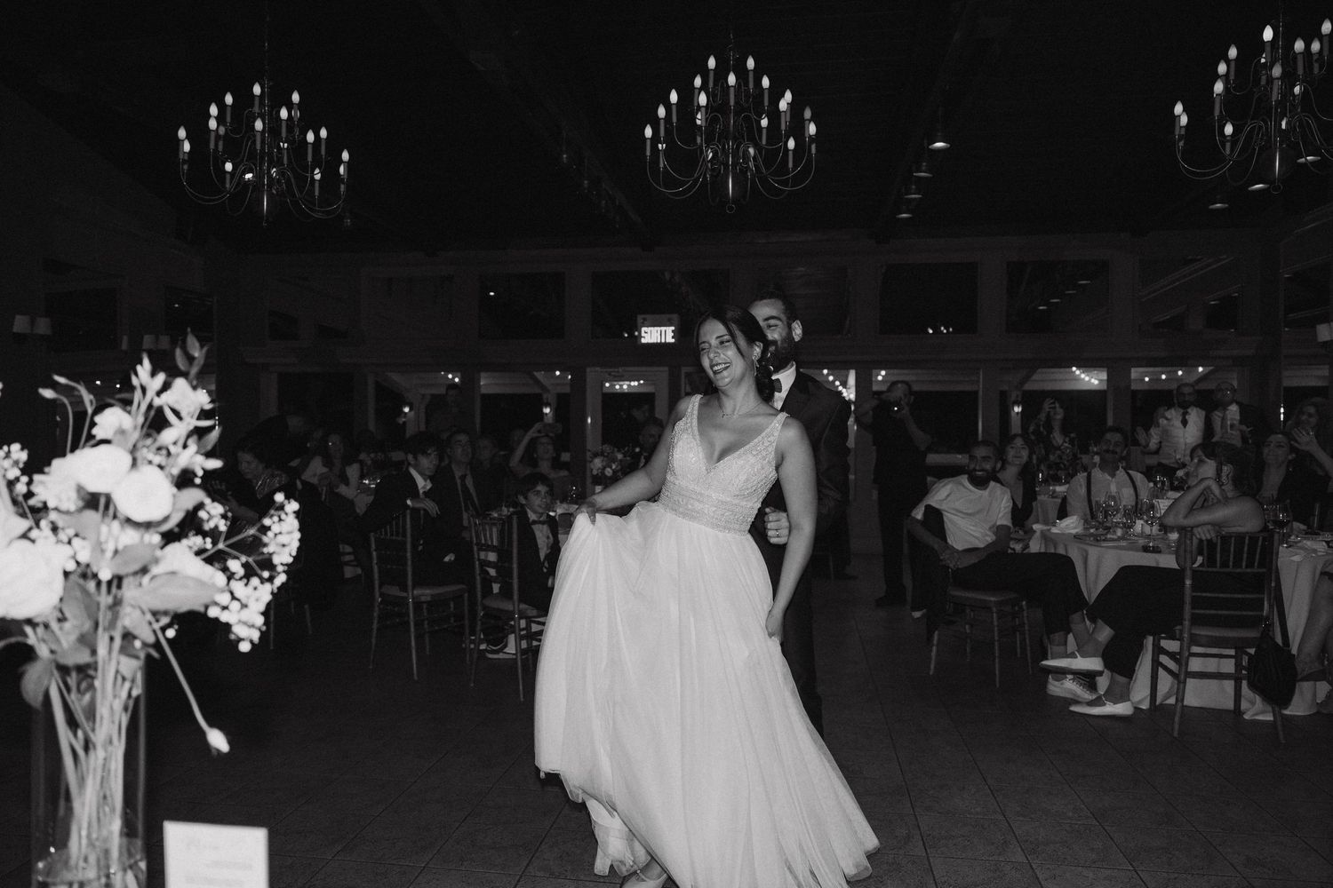 Black and white photo of wedding reception with couple dancing under chandeliers.