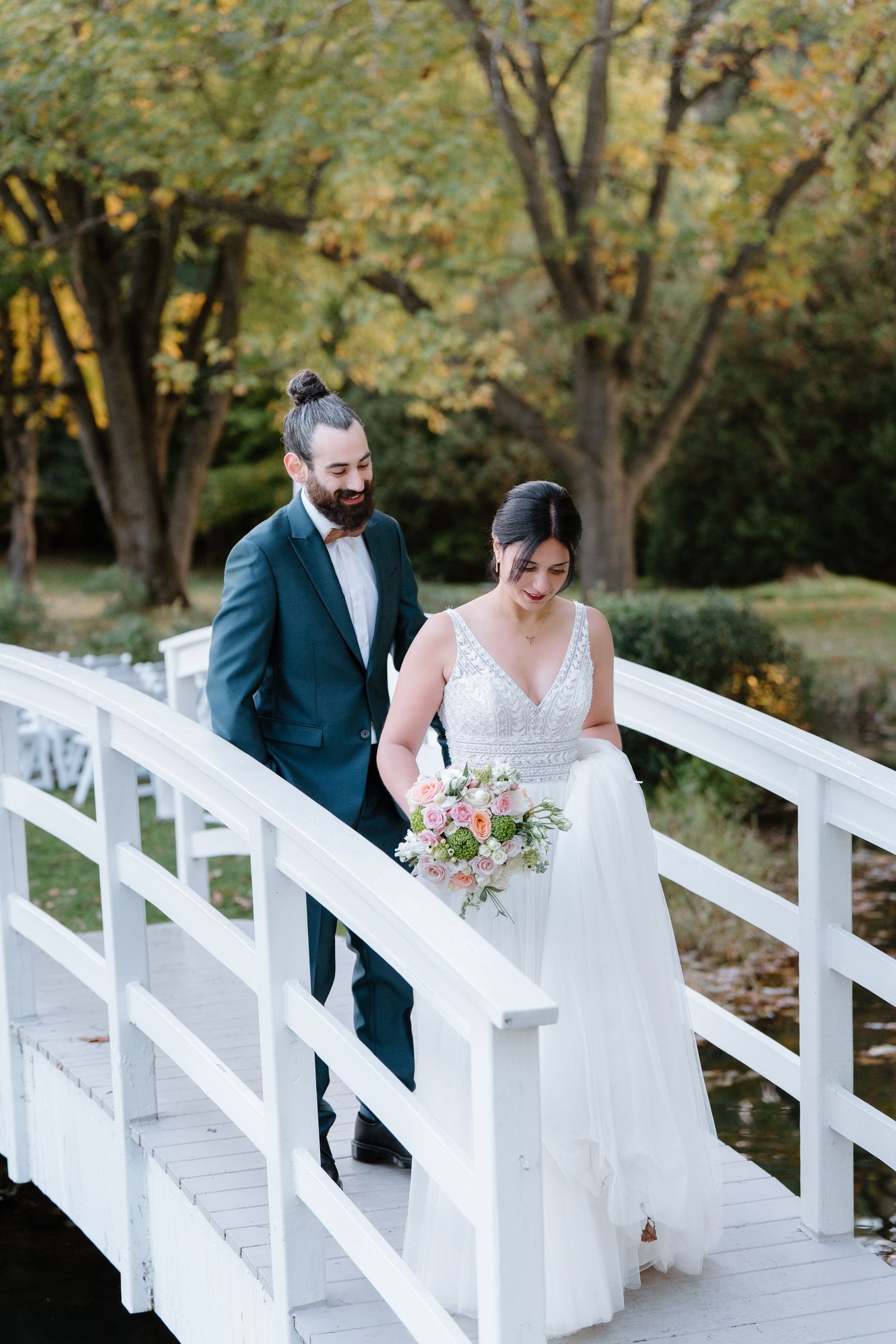 Wedding couple walking across a white wooden bridge surrounded by autumn trees.