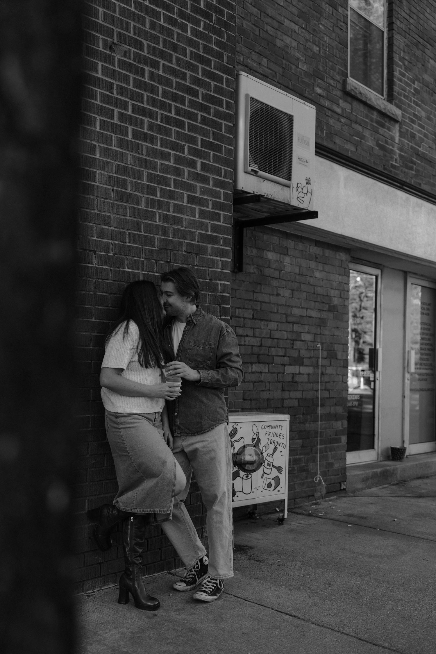 Black and white photo of a couple sharing an intimate moment against a brick wall on a city sidewalk.