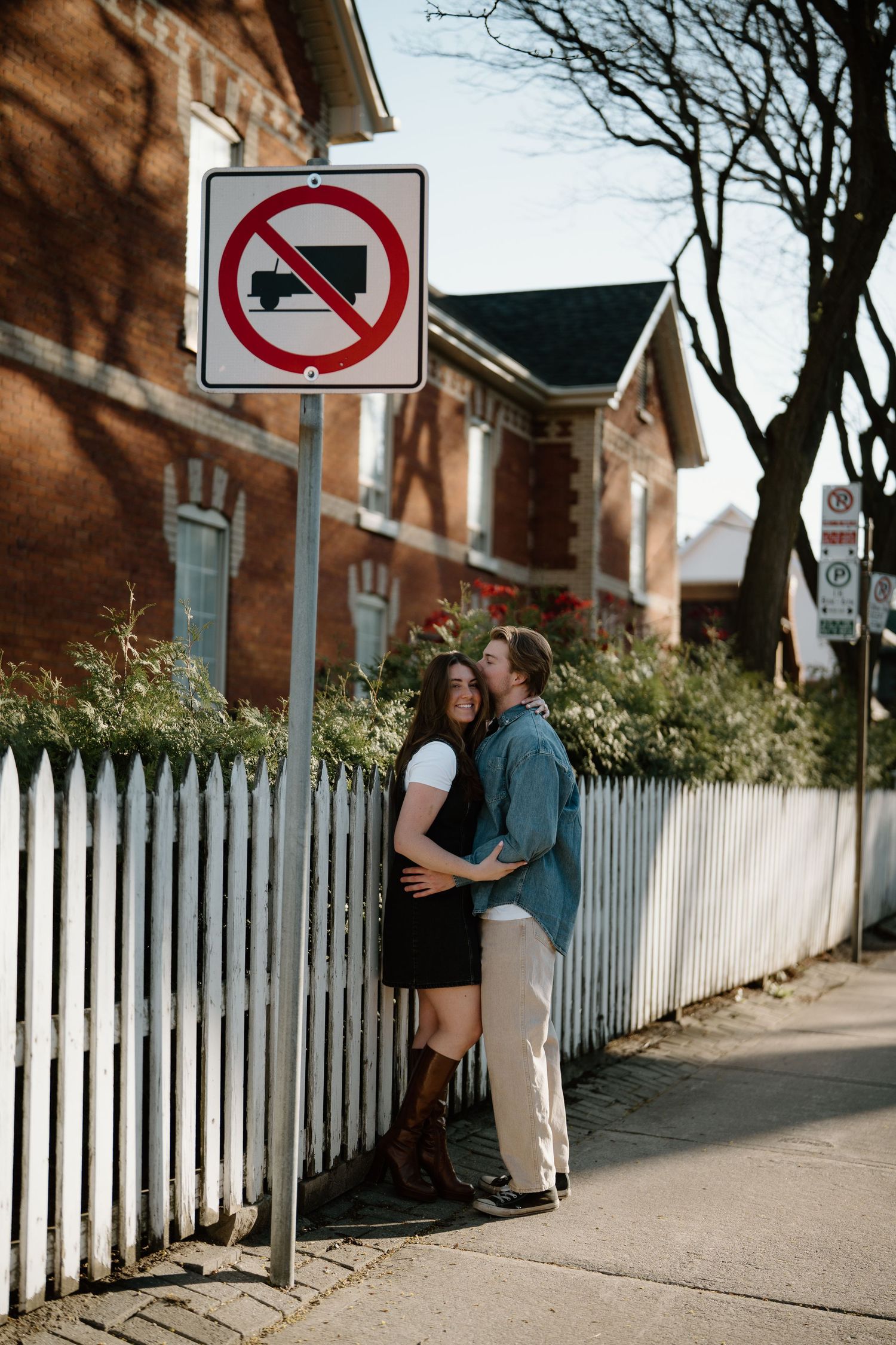 Couple stands intimately by white picket fence near brick buildings with no trucks sign above them on residential street.