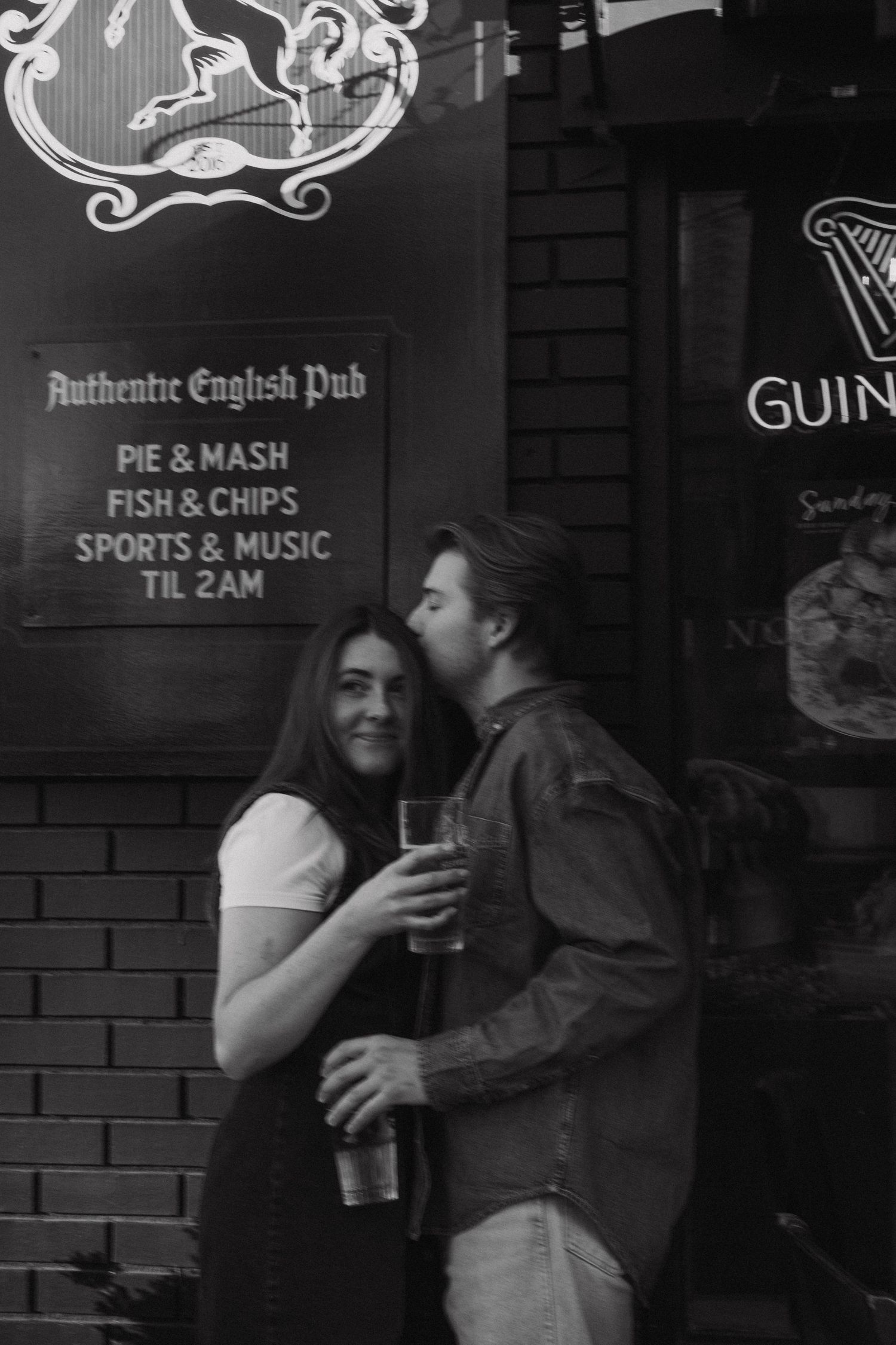 A couple sharing an intimate moment outside an authentic English pub with Guinness signage in black and white photography.