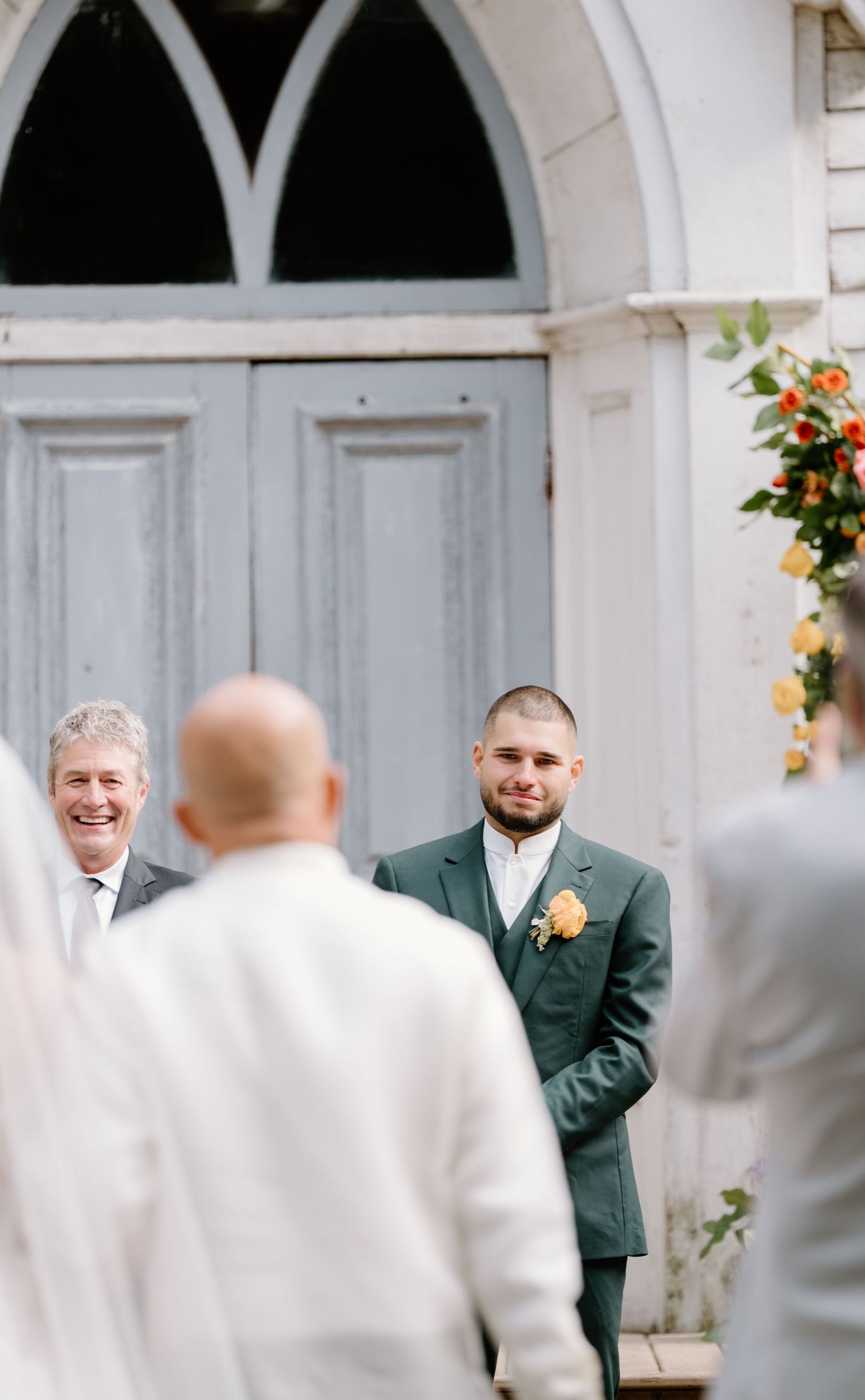 Wedding party gathers outside a white church with an arched doorway during a sunny outdoor ceremony.