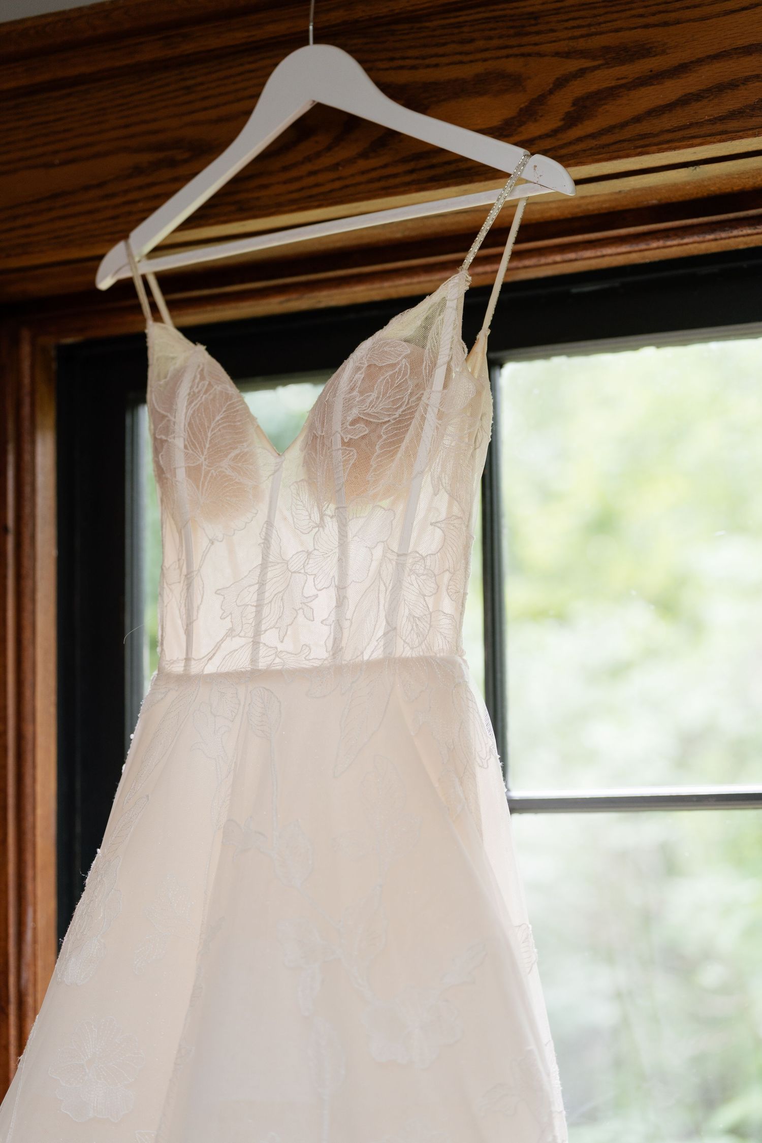 A delicate white wedding dress with lace bodice and spaghetti straps hangs on a white hanger by a window.