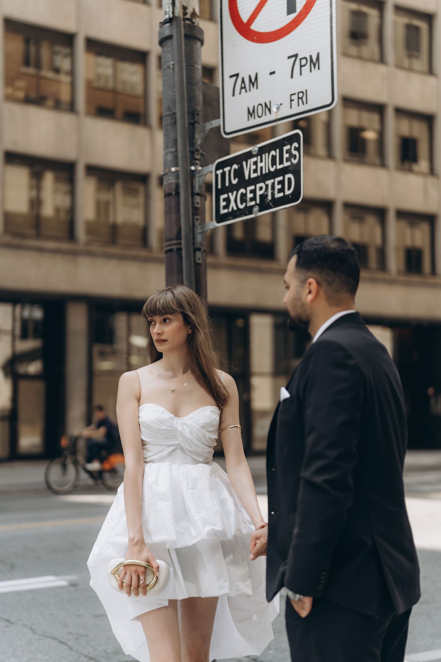Couple in formal wedding attire walking on urban city sidewalk with parking signs visible in background.