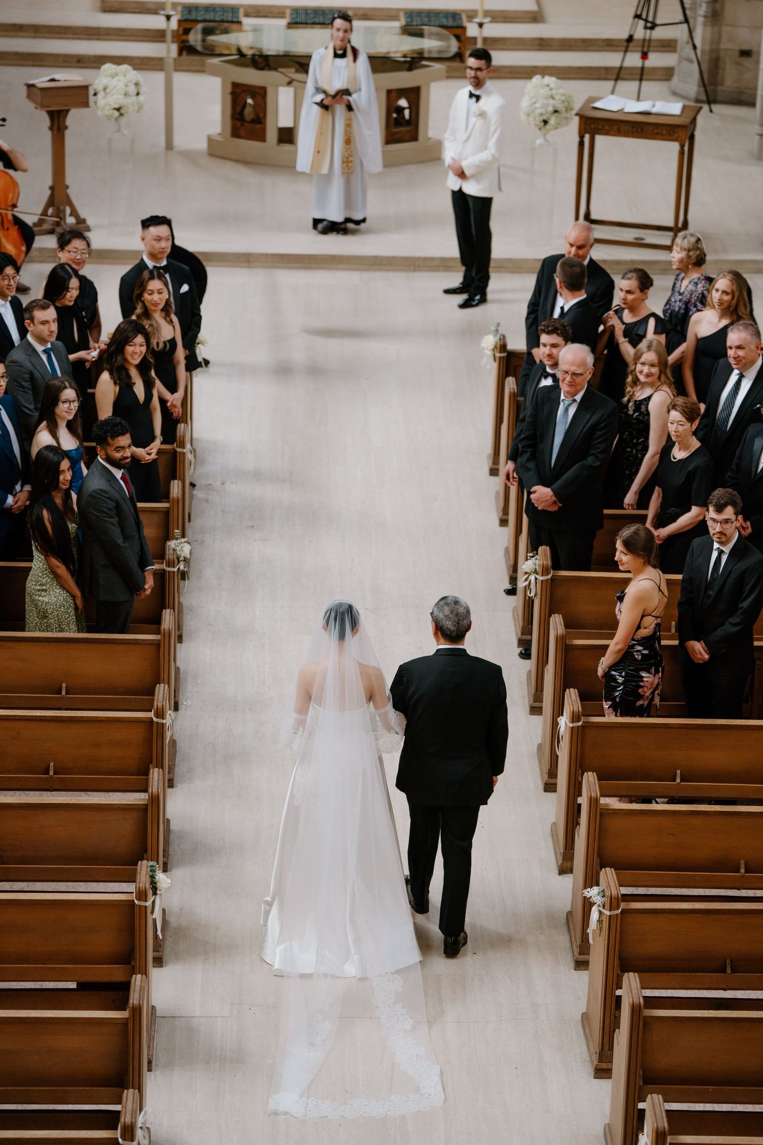 A wedding ceremony in progress showing a bride walking down the church aisle with her escort as guests look on from the pews.