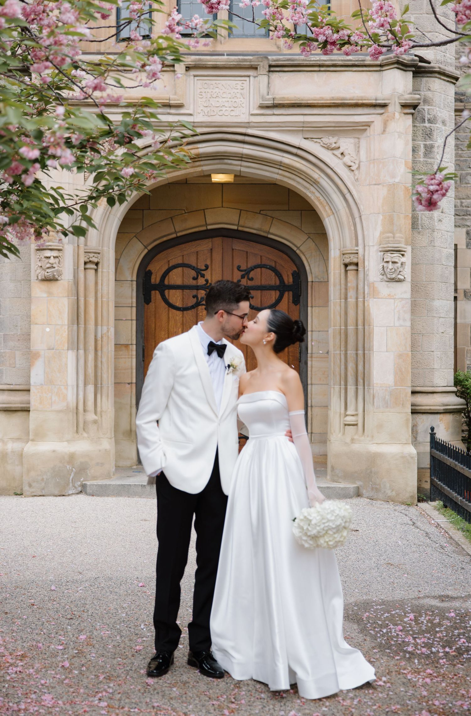 Elegant couple shares a romantic moment in front of ornate stone archway with pink cherry blossoms blooming nearby.