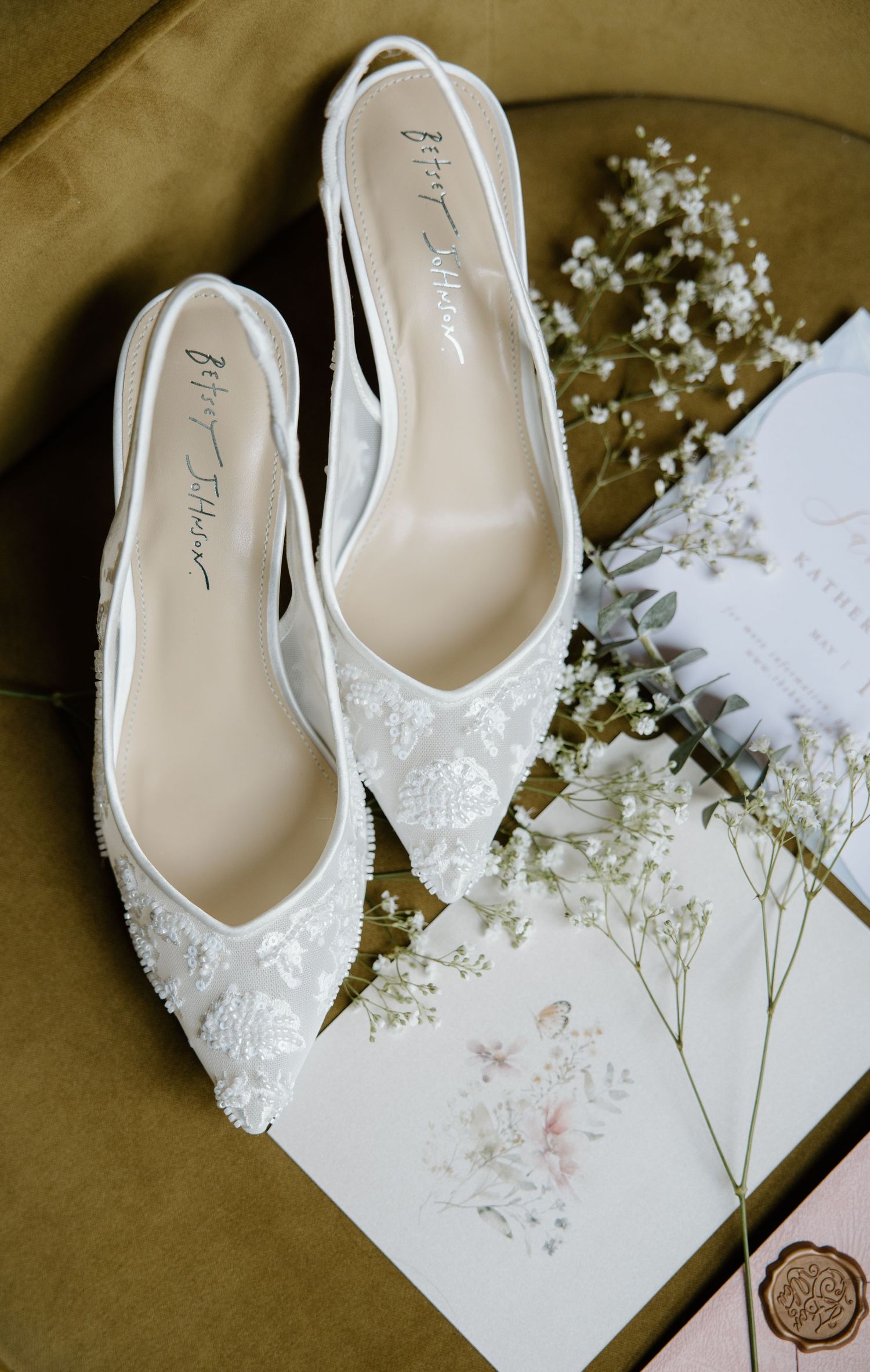 Elegant white lace bridal flats displayed with delicate baby's breath flowers and a floral invitation card on neutral background.