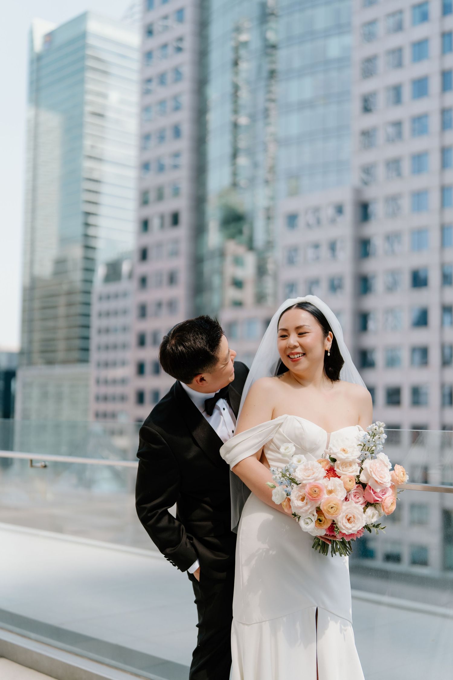 A couple in wedding attire stand on a rooftop terrace with modern city buildings in the background.
