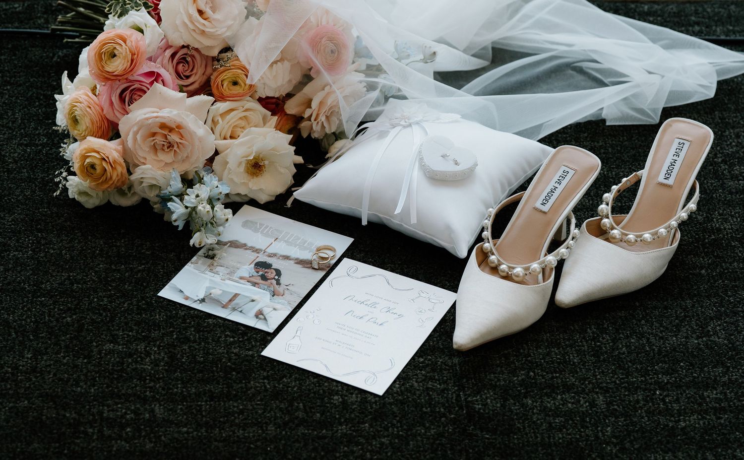 Beautiful wedding details with peach and pink roses, white invitation cards, and studded white heels on dark surface.