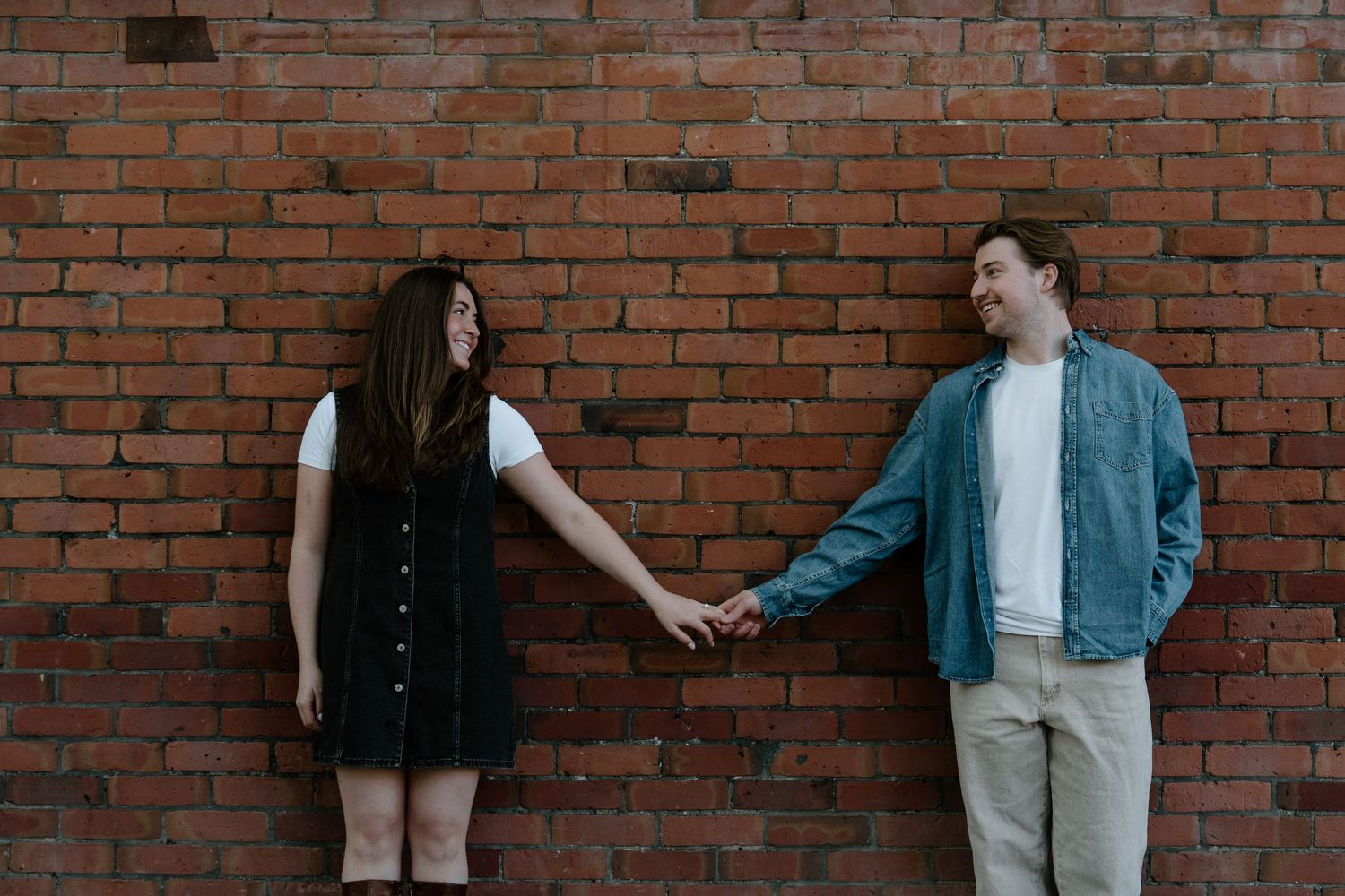 Two people holding hands and standing against a rustic brick wall, with one wearing a black dress and another in denim.