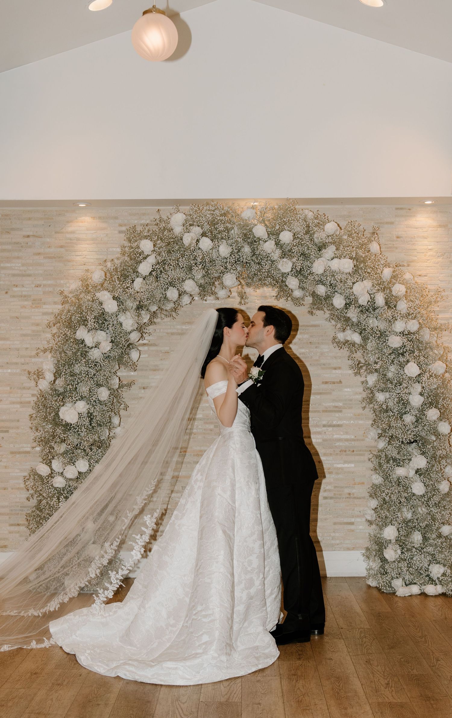 Wedding couple poses in front of elegant white floral arch backdrop in indoor venue during romantic photoshoot.
