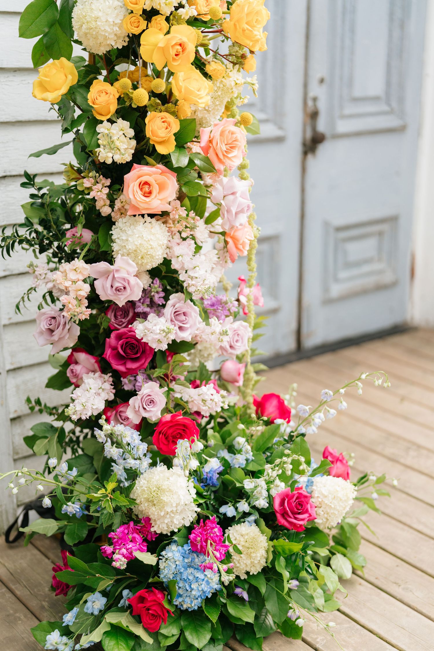 Elegant floral arrangement featuring roses in yellow, pink and red with white hydrangeas cascading down against a white door.