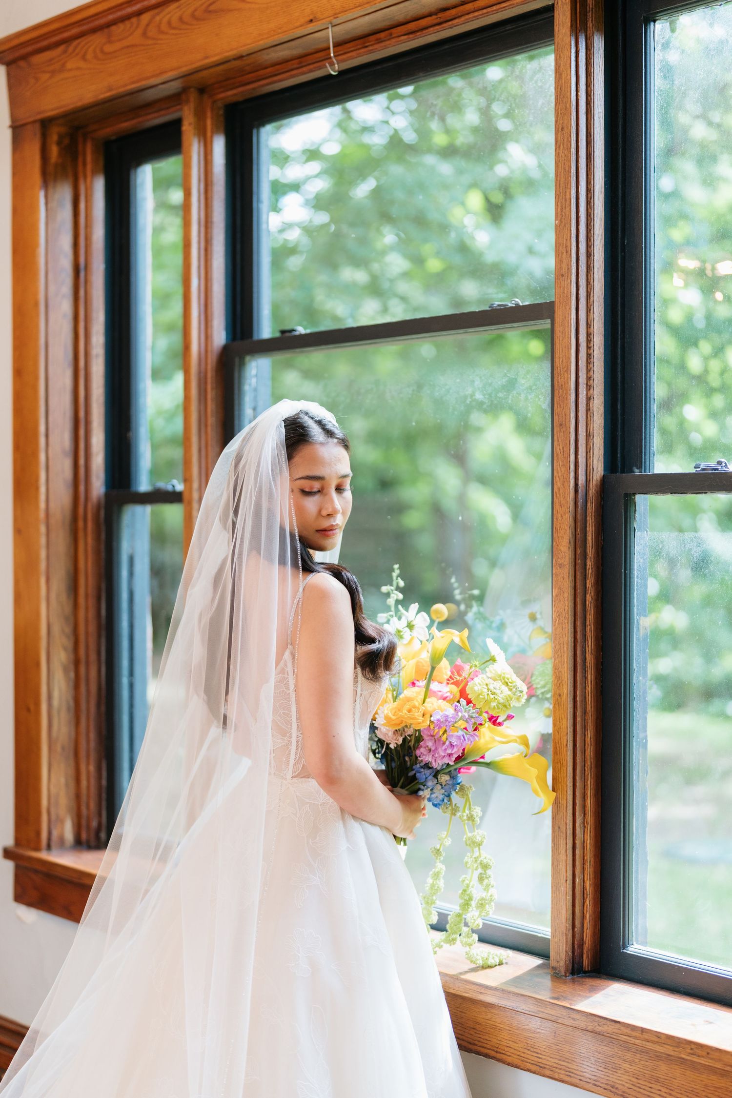 A bride in a white wedding gown and veil poses by large windows in a room with wooden trim.