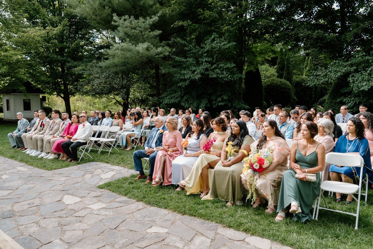 Wedding guests seated on white folding chairs outdoors during a garden ceremony.