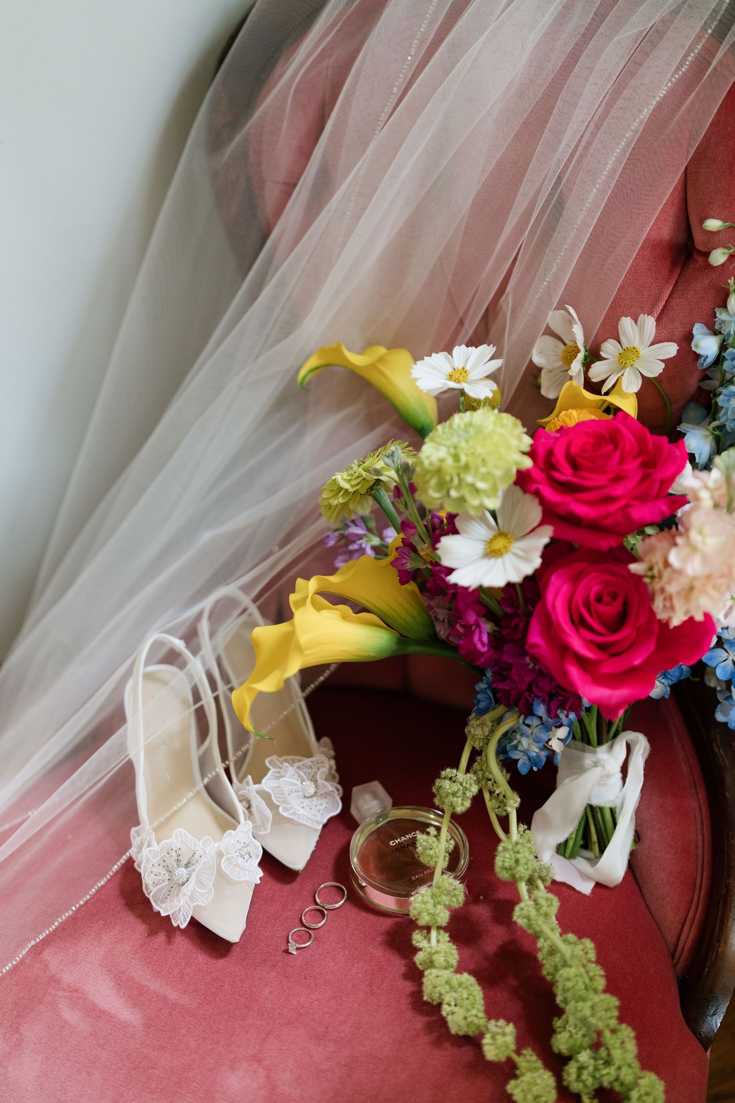 Wedding accessories including white lace shoes, rings, and a colorful bouquet with roses and daisies on pink fabric.
