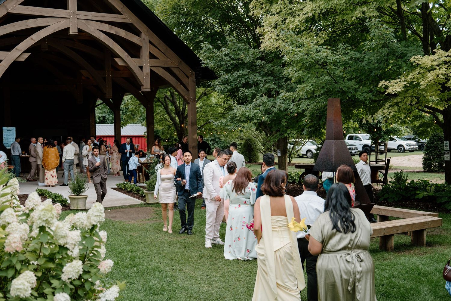A group gathers outdoors under trees for a casual summer celebration with picnic tables and benches.