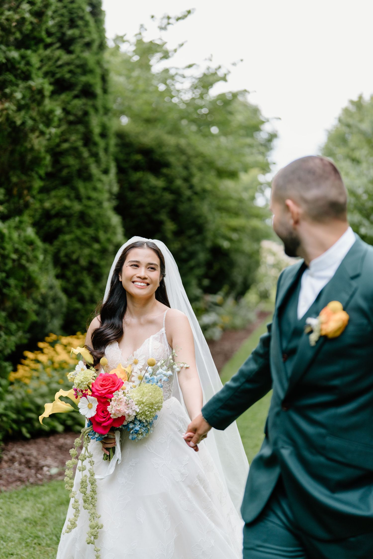 A couple in wedding attire stands in a garden setting with colorful flowers and greenery.