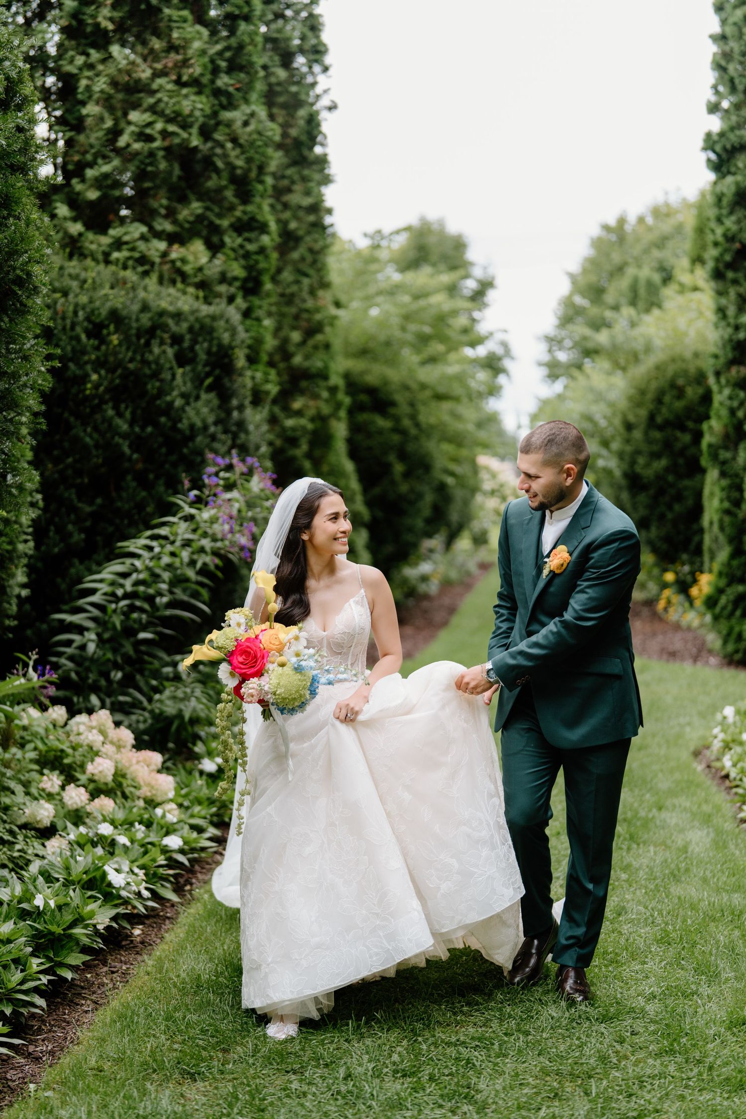 Wedding couple strolls along manicured garden path with towering evergreens and blooming flowers.