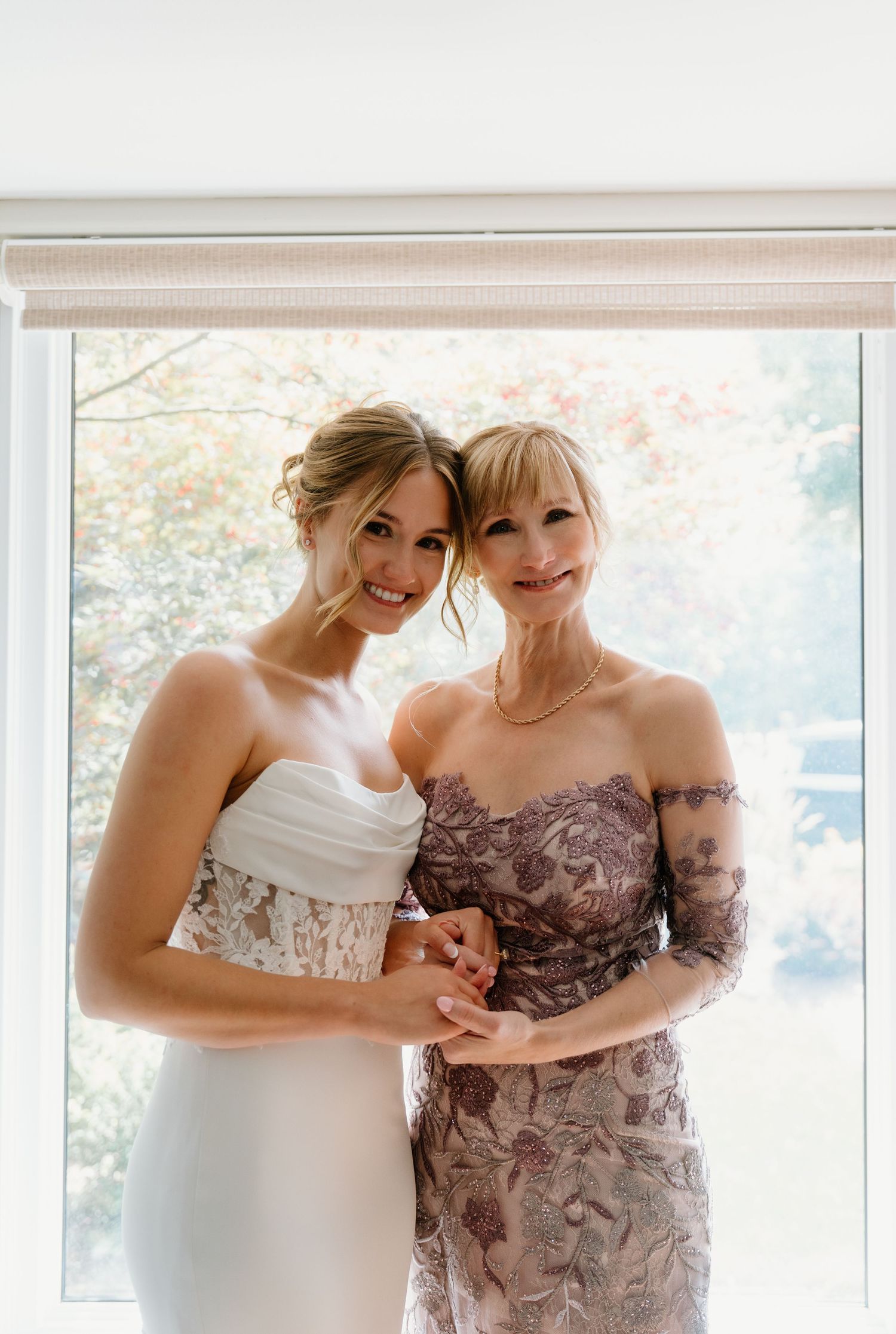 Two people in formal attire share a tender moment while standing by a bright window.
