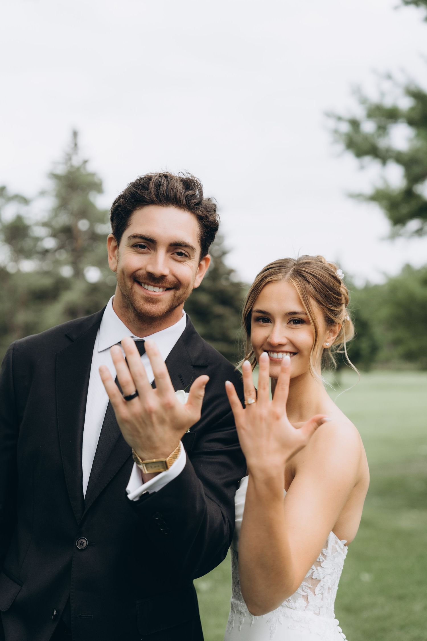 A newly married couple showing off their wedding rings while smiling outdoors in a grassy field.