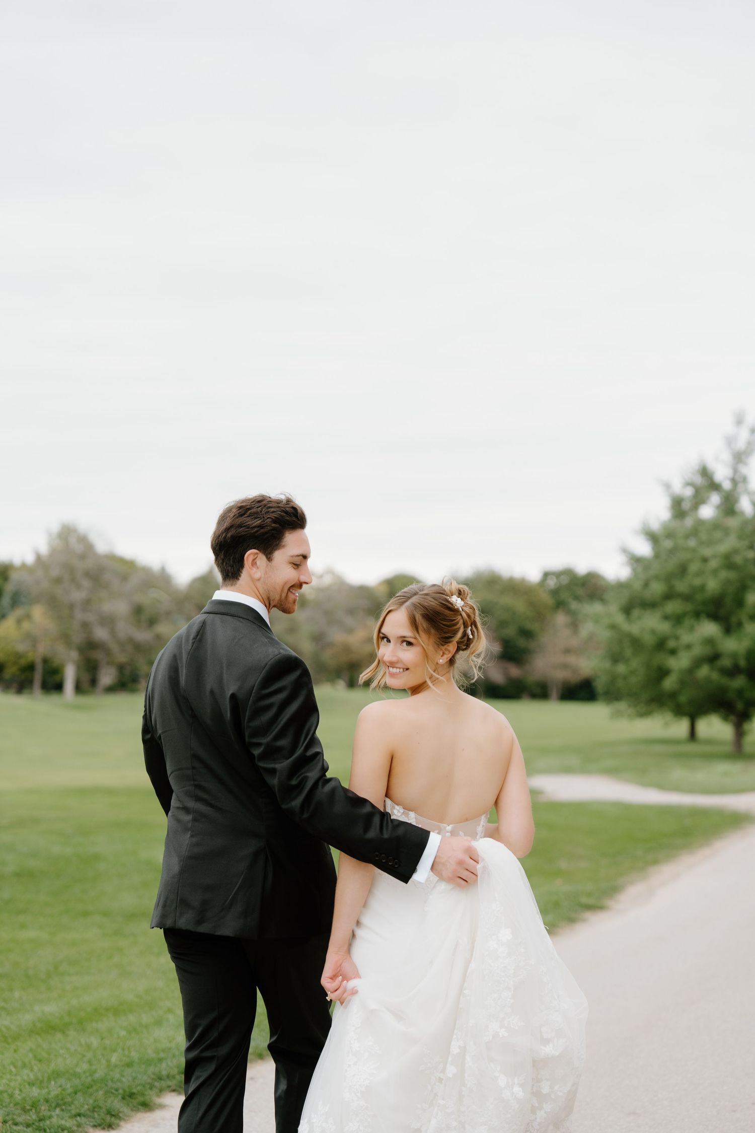 A joyful wedding couple walks together along a winding path through a scenic green park.