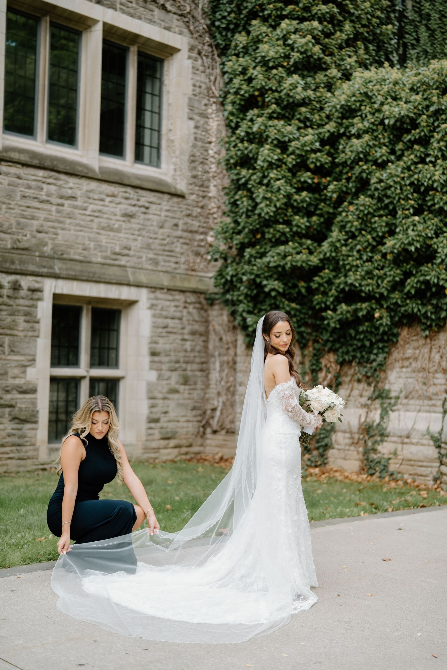 A wedding scene outside a stone building with a long flowing veil being arranged on a sunny day.