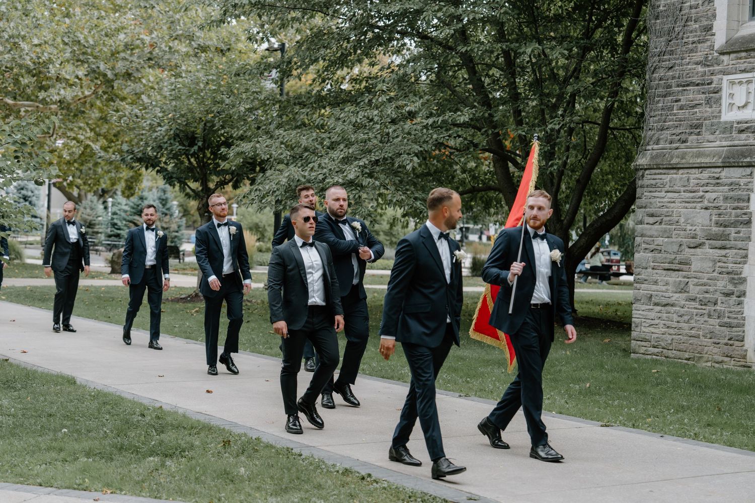 A group of people in formal black tuxedos walk together on a path near a stone building while one carries a red flag.