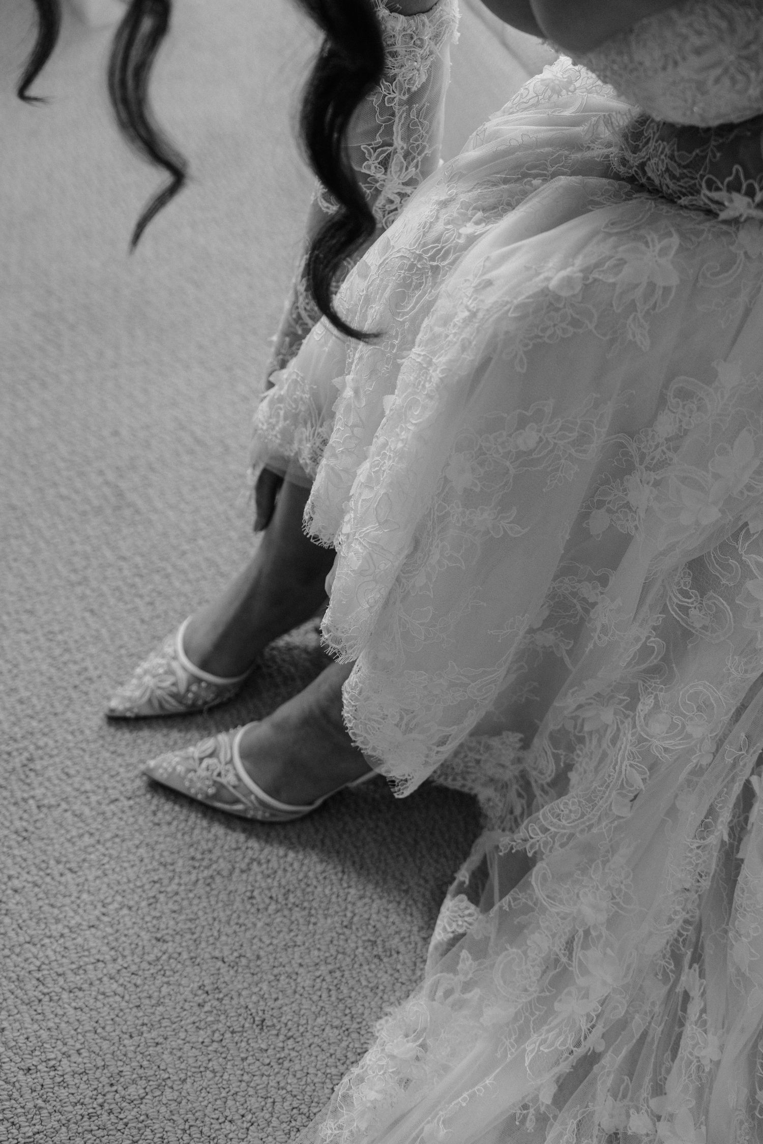 Black and white detail of ornate bridal shoes and lace wedding dress hem on carpet.