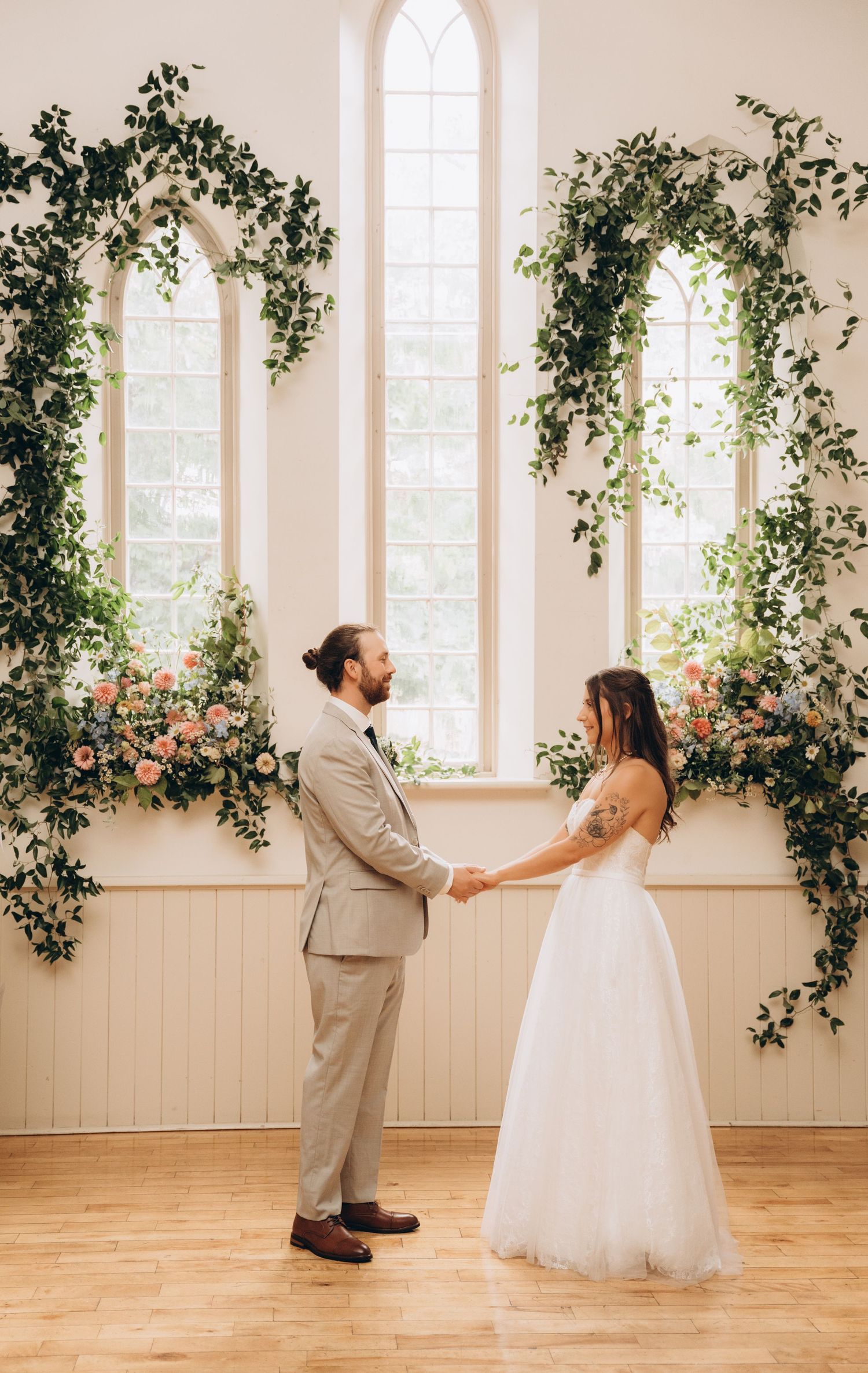 A couple in wedding attire playfully dances and spins across a bright sunlit room with floral window decorations.