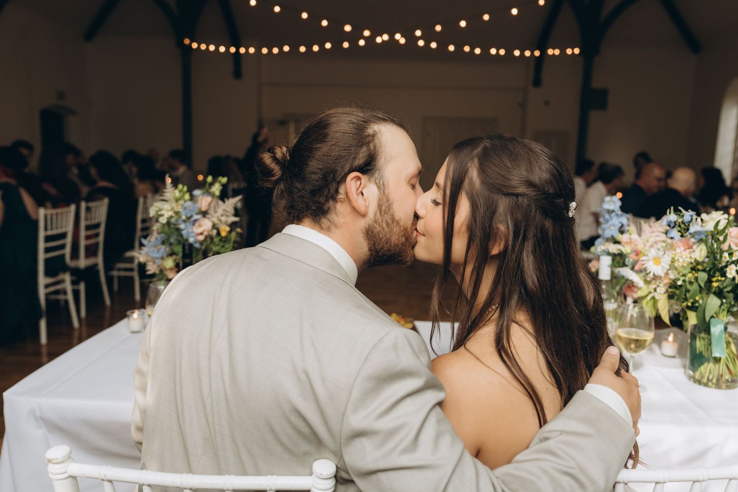 A romantic couple shares a kiss at a wedding reception under twinkling string lights and floral decorations.