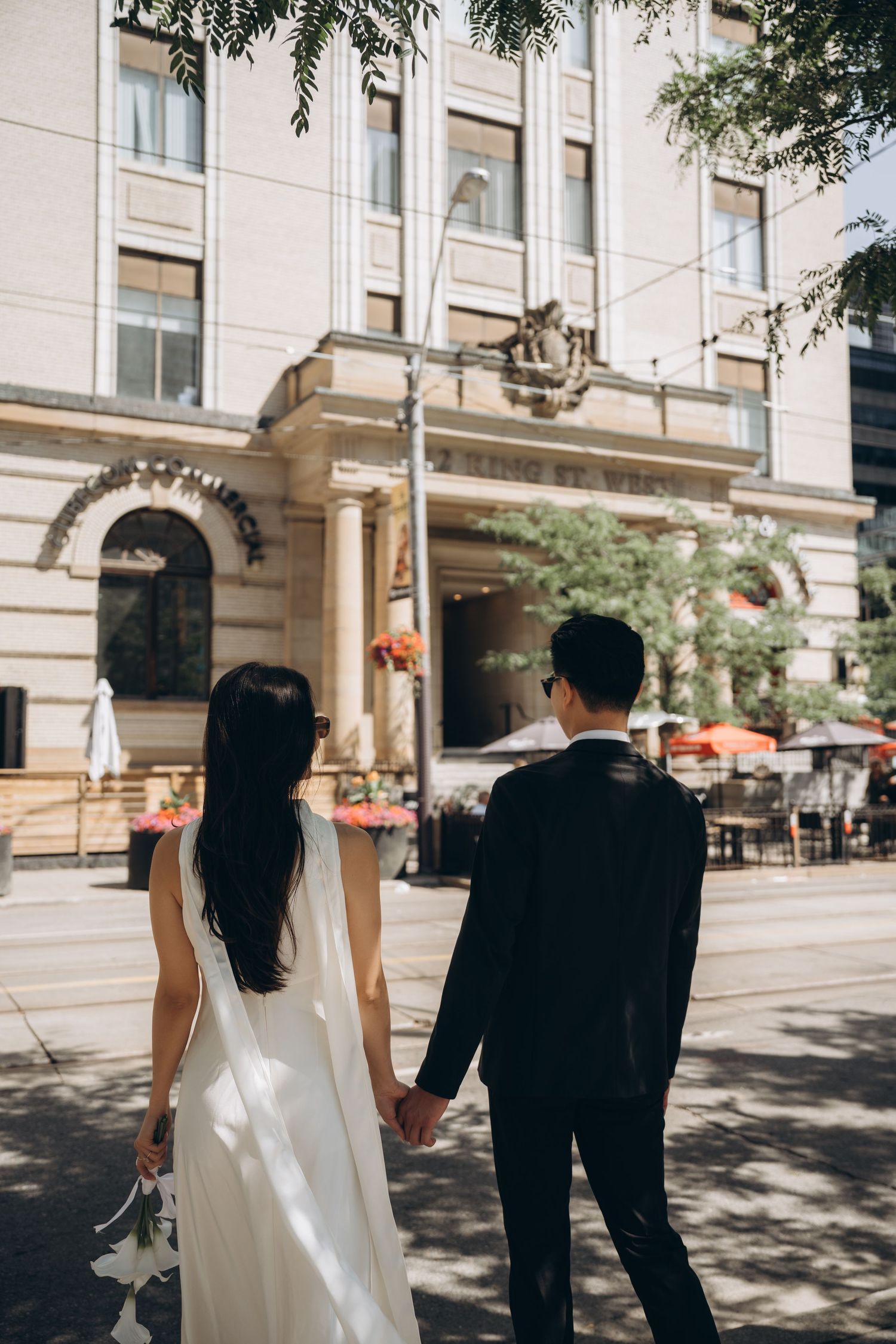 A couple in wedding attire holding hands while walking on a sidewalk near an elegant historic building facade.