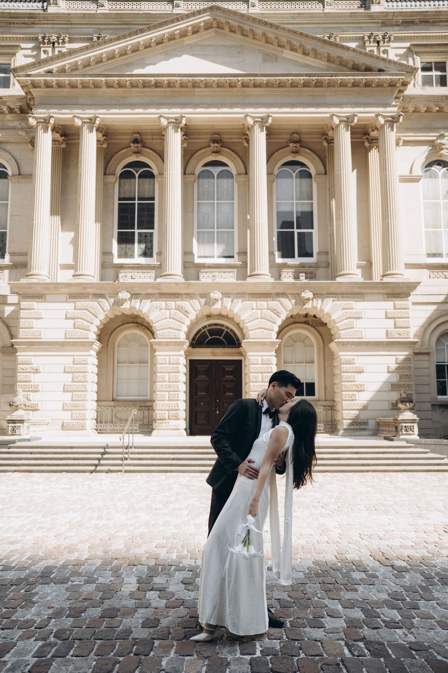 A romantic couple embraces in front of a grand neoclassical building with ornate columns and architectural details.