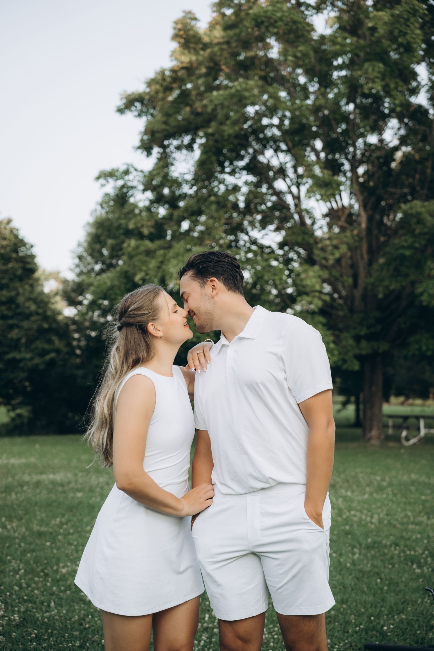 Couple wearing matching white outfits sharing an intimate moment in a park setting with green trees in background.