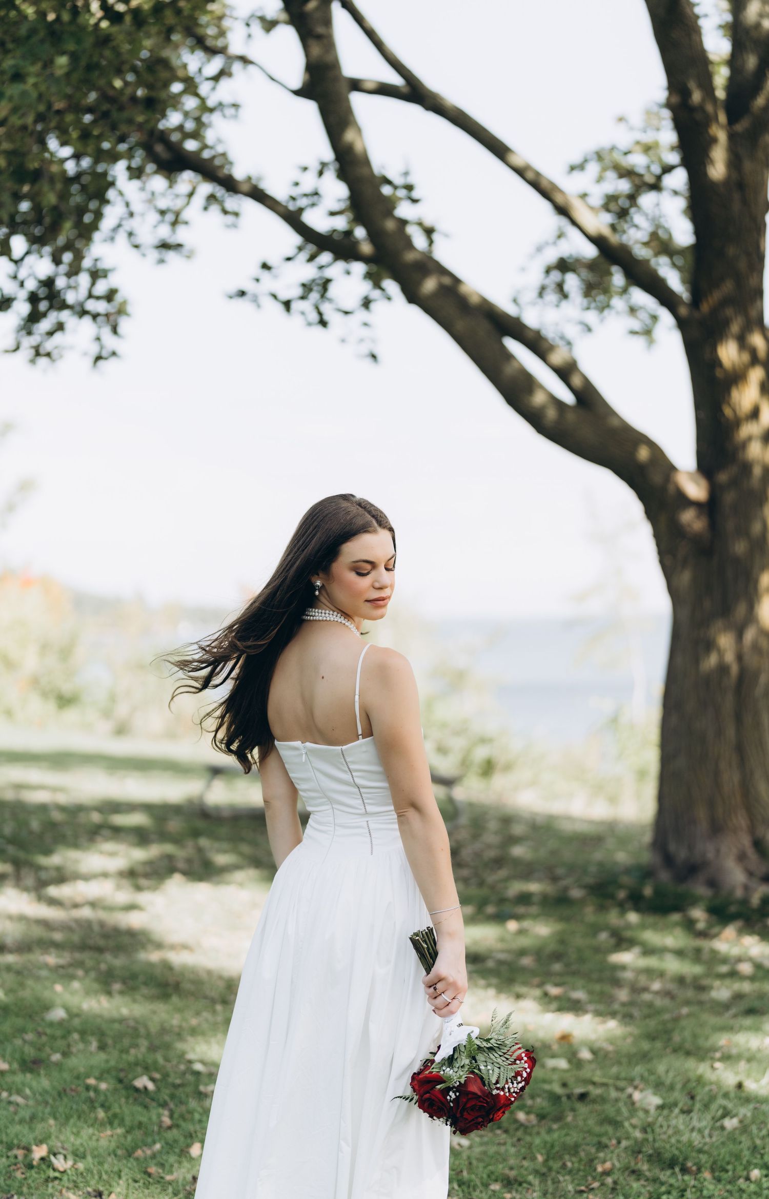 A person in a flowing white wedding dress stands among trees in a sunny outdoor setting holding a burgundy bouquet.