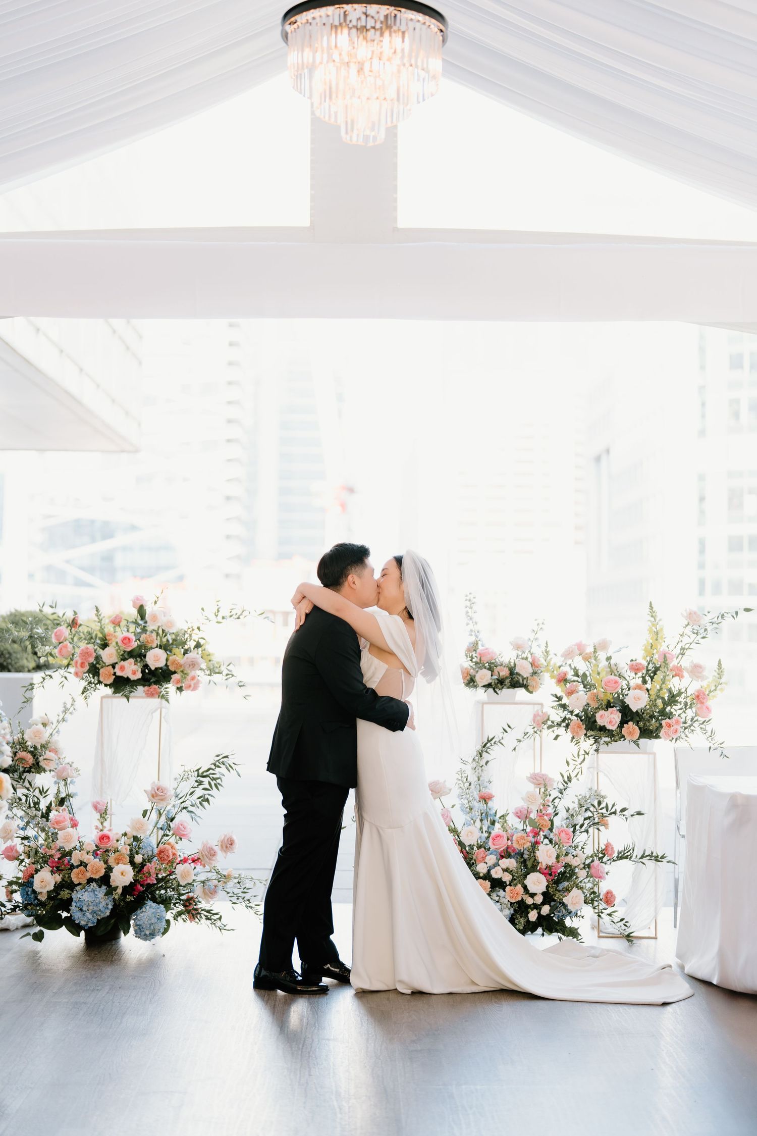 An elegant ballroom wedding ceremony with crystal chandelier and romantic floral arrangements in pink and white tones.