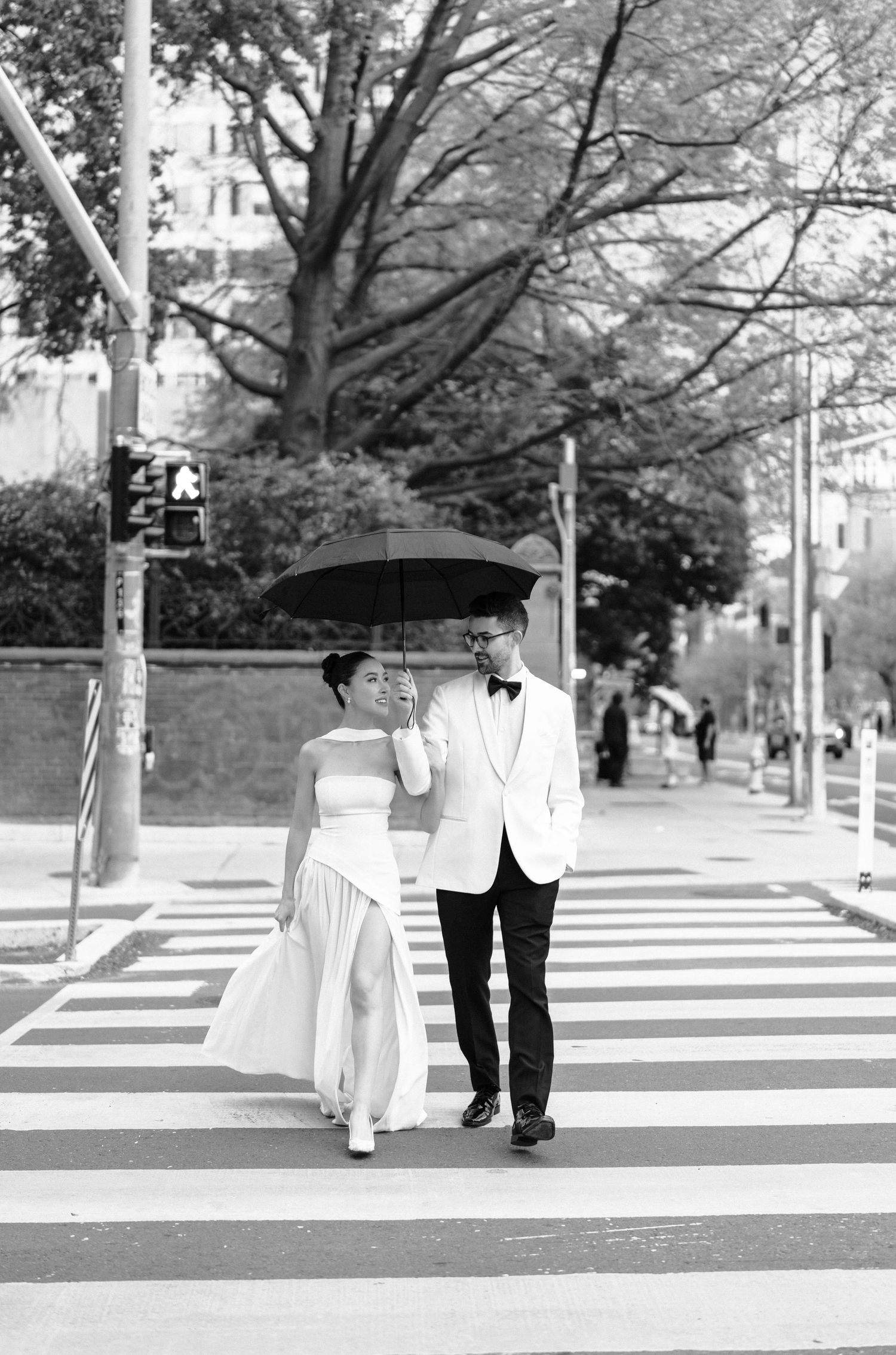 Black and white composite photo of two figures walking across a crosswalk with blooming trees in the background.