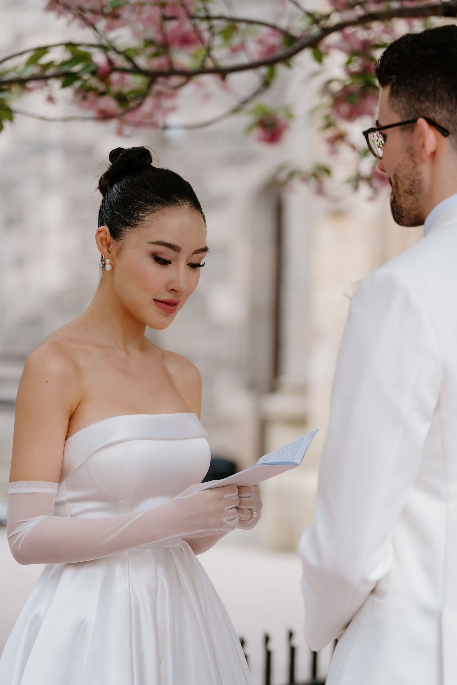 A couple in wedding attire stands under cherry blossoms as the bride reads from a paper during their outdoor ceremony.
