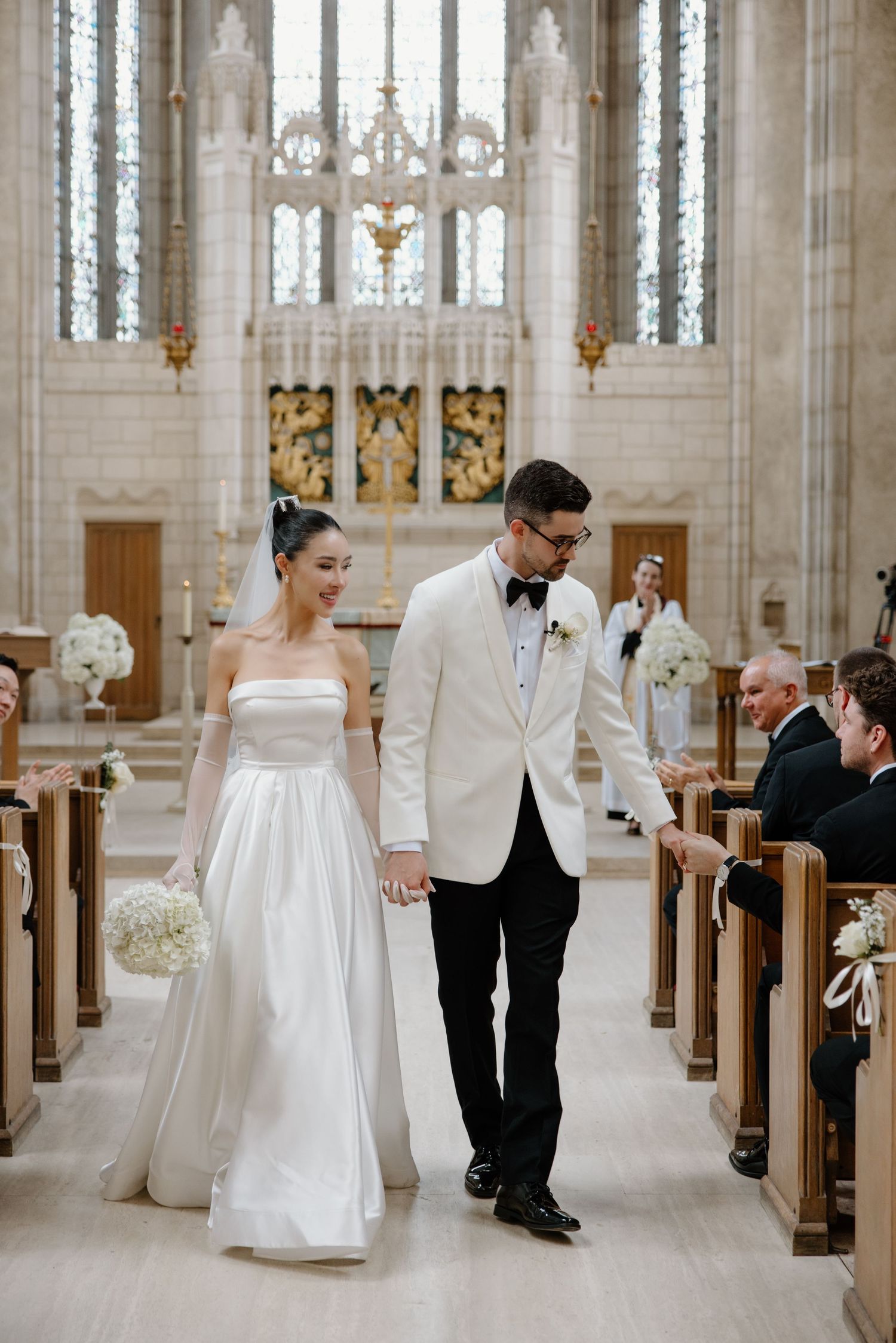 A couple walks down the aisle in a sunlit gothic church with stone columns and stained glass windows during their wedding ceremony.