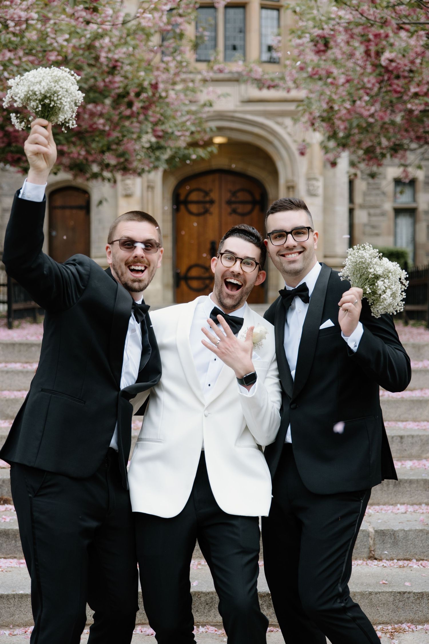 Three groomsmen in black and white tuxedos celebrate with flowers in front of stone building with cherry blossoms.
