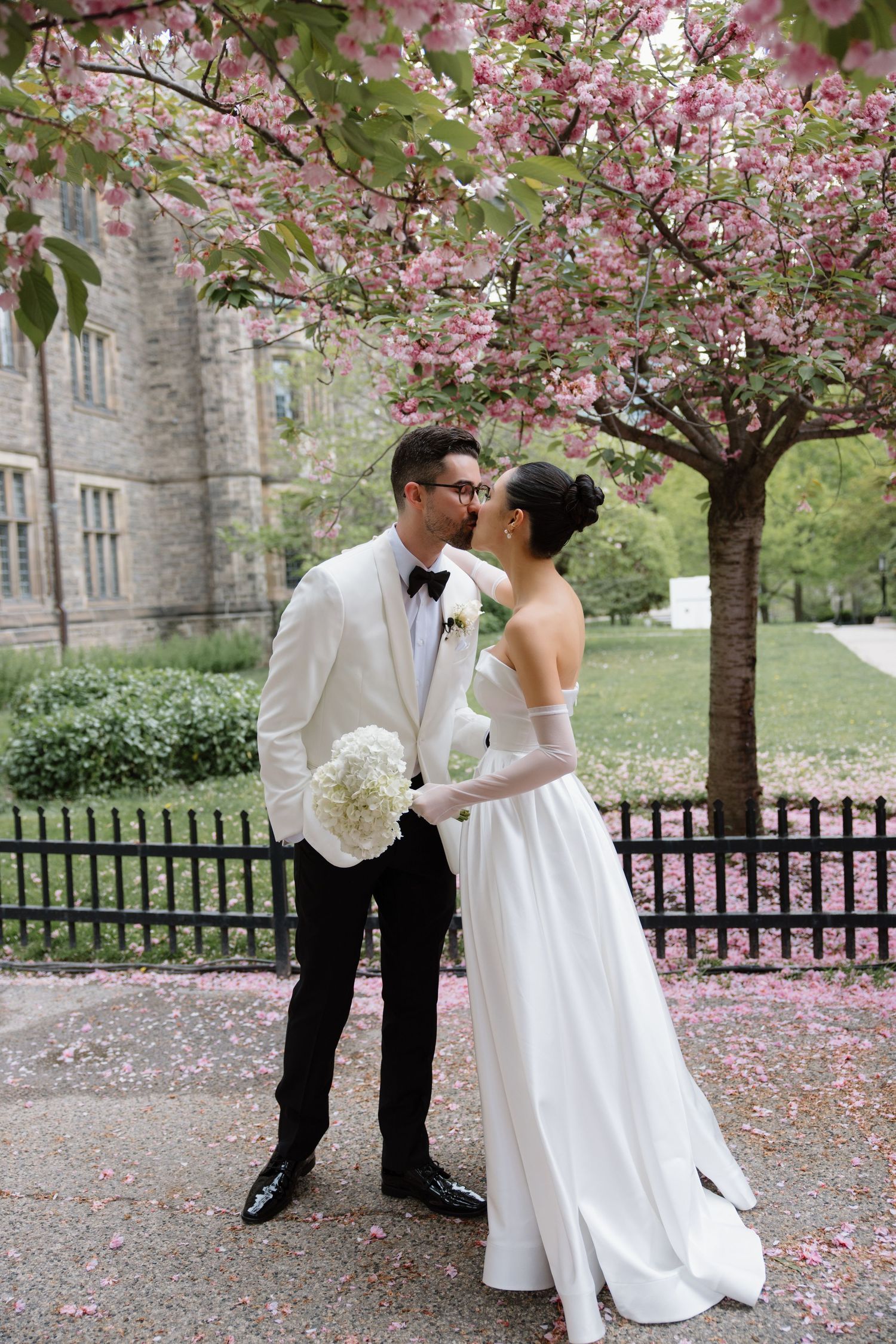 A couple in wedding attire embraces under blooming cherry blossoms near a historic stone building in springtime.