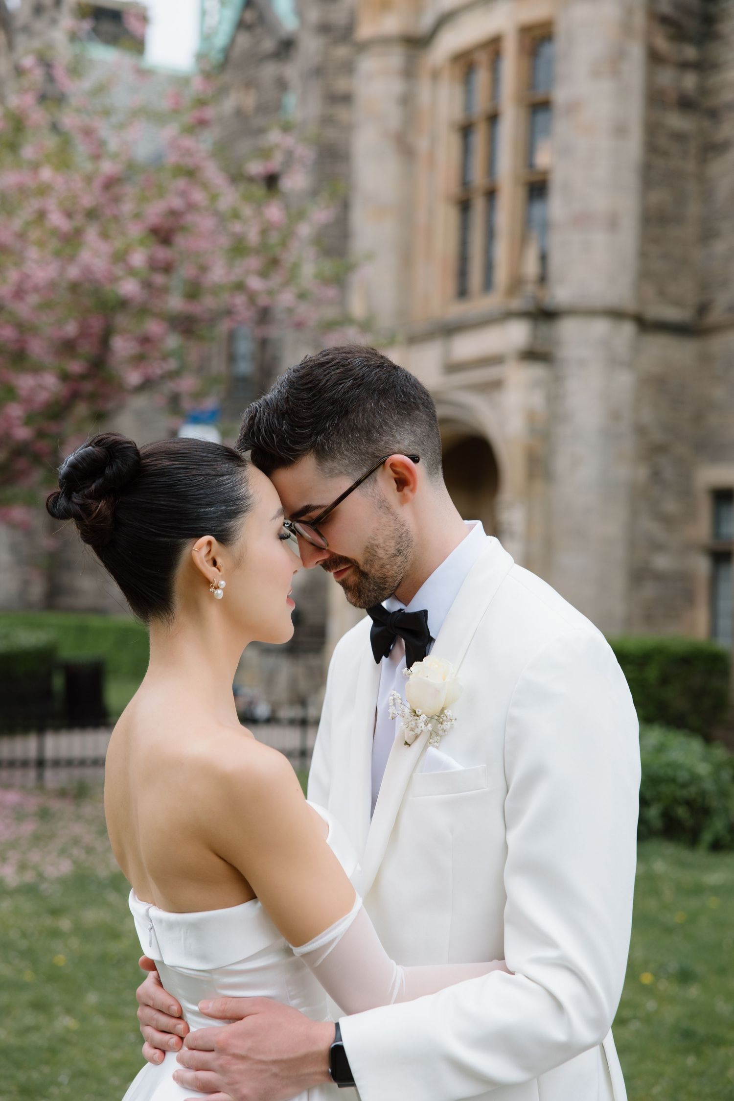 A romantic moment between newlyweds in formal attire standing by a gothic stone building with blooming spring trees.