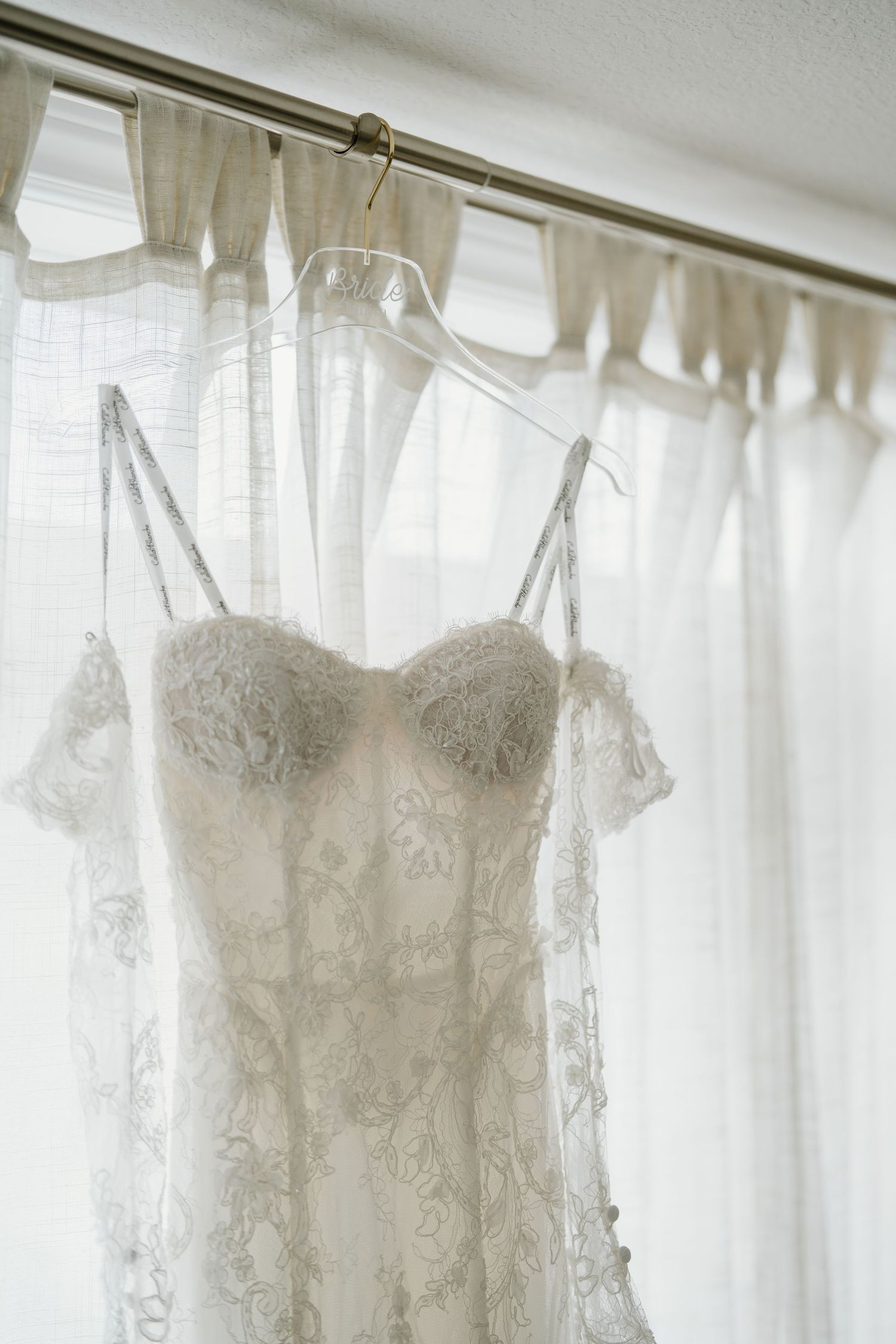 White wedding dresses hanging on display with delicate lace details and soft lighting through sheer curtains.